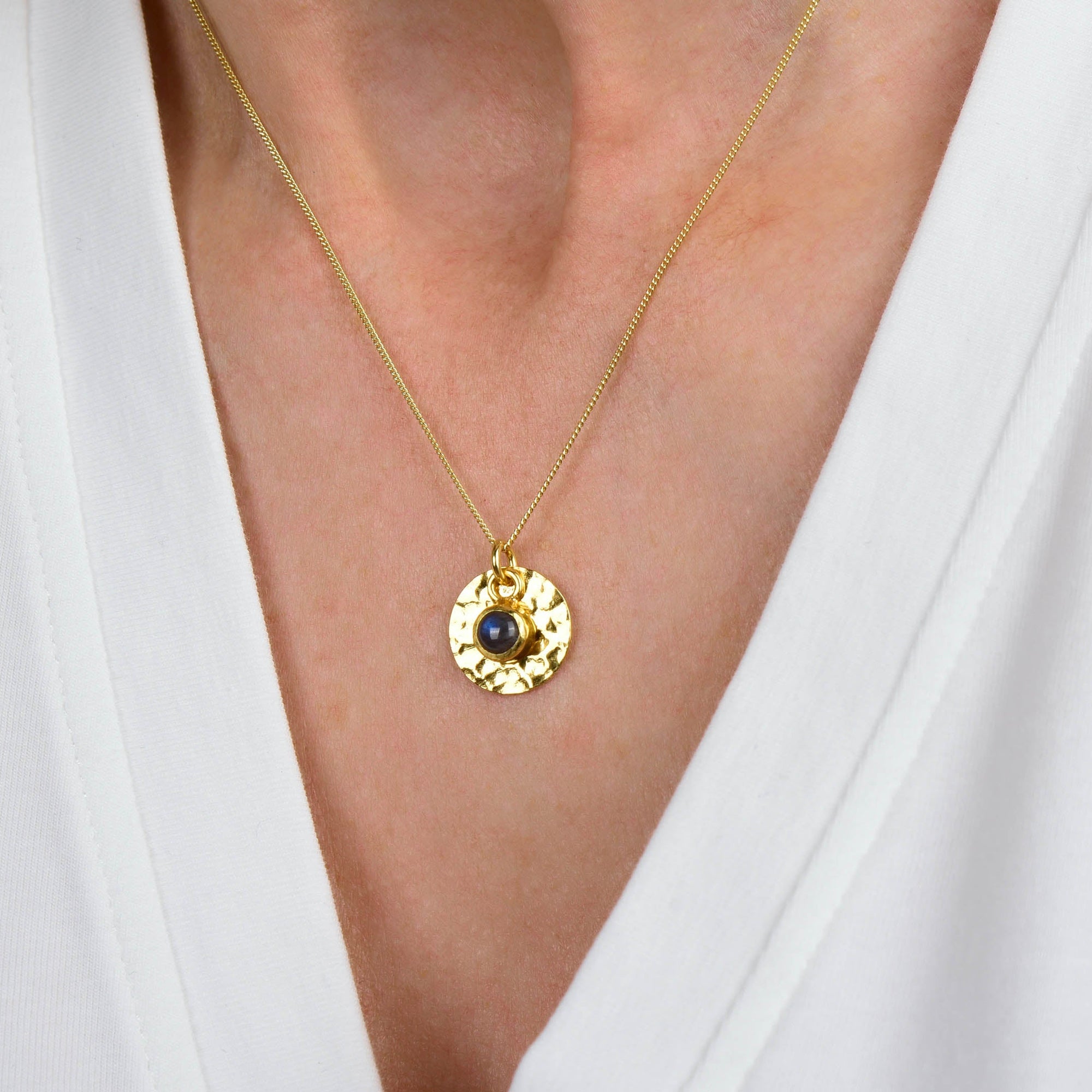 A Labradorite Luna Disc Necklace, featuring a gold pendant with a dark blue labradorite stone, is worn around a person's neck and is visible against a white V-neck shirt.