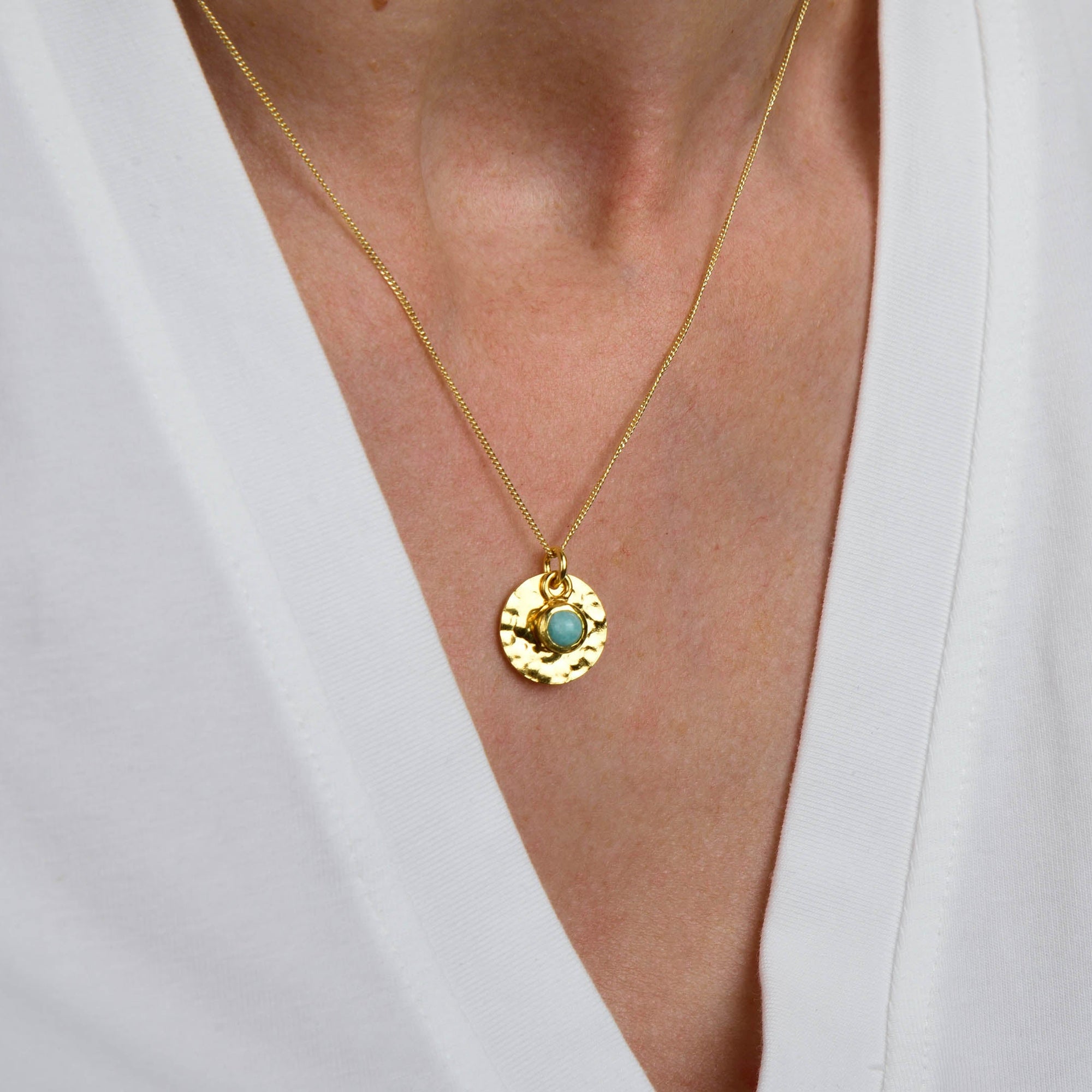 A person wearing a white V-neck shirt and the Amazonite Luna Disc Necklace, which features a gold plated, round hammered disc pendant with an amazonite stone in the center.