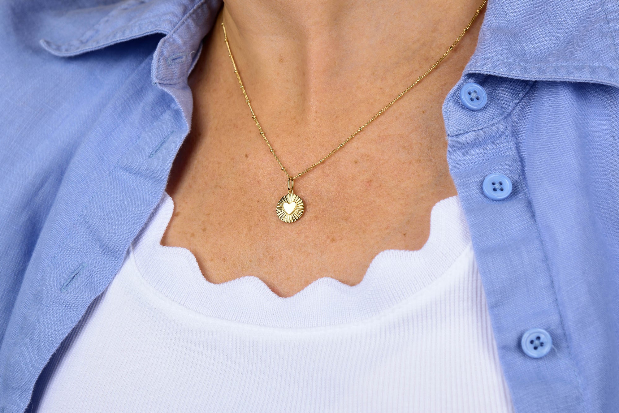 A person wearing the Shining Heart Necklace Sterling Silver layered with a gold sunburst pendant over a white scalloped-edge top and a light blue button-up shirt.