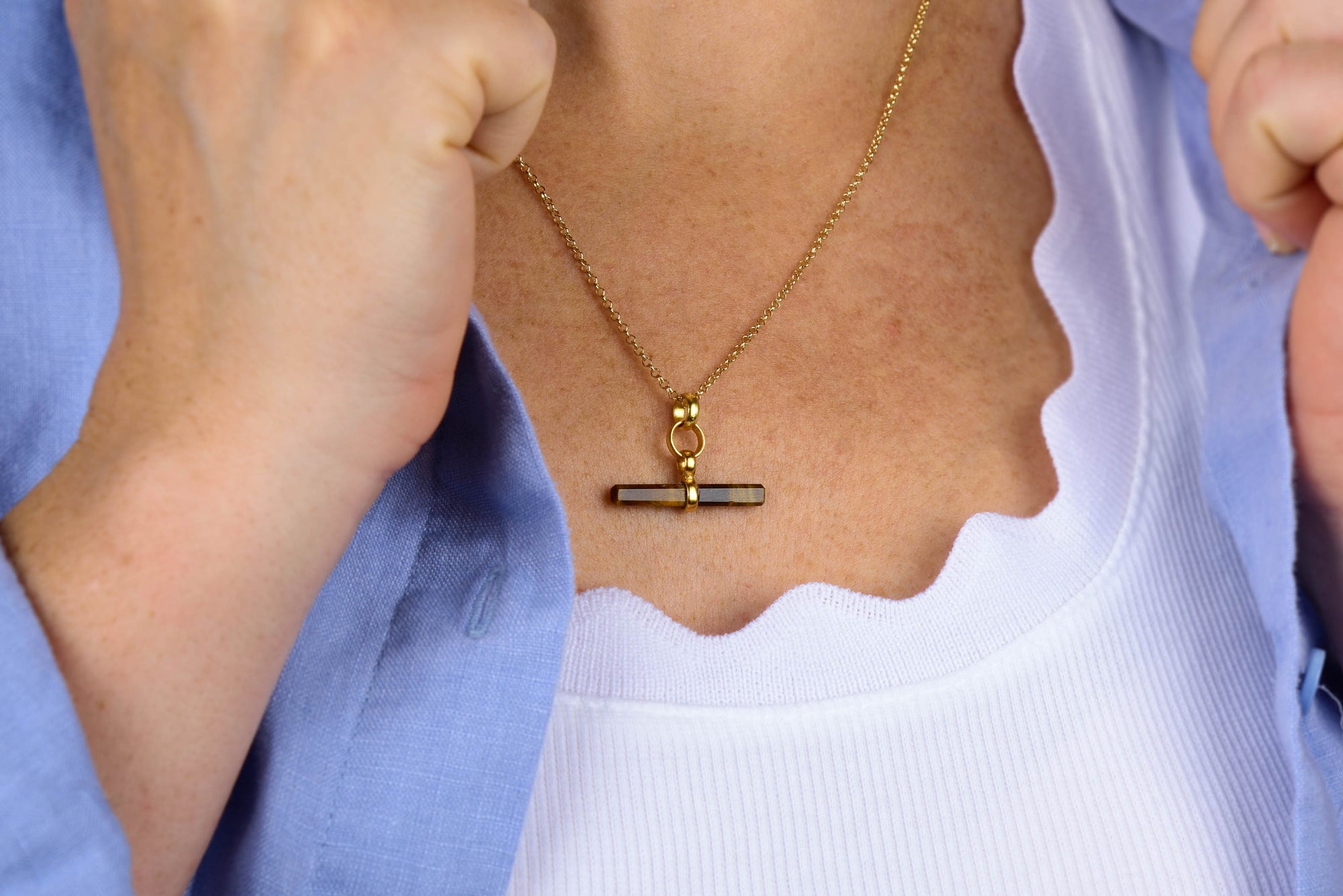 A person wearing a white scalloped top and blue shirt displays the Tiger Eye T-Bar Necklace, featuring a gold T-bar pendant with a horizontal bar detail.