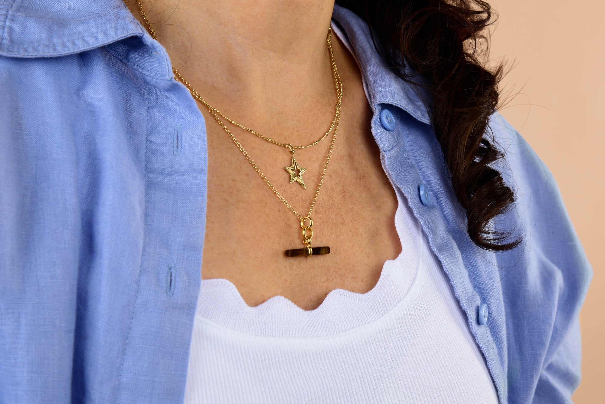 A woman wearing a blue button-up shirt over a white scallop-edged top, adorned with two gold necklaces—one featuring the Stellar Starburst Necklace and the other with a simple bar pendant.
