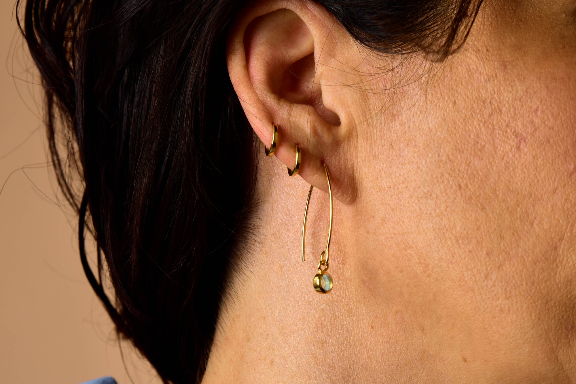 Close-up of a person's ear with three small gold hoop earrings in the upper ear and one large gold hoop earring with a green bead in the lower lobe. The person has short, dark hair and light skin. The striking arrangement is complemented by the subtle elegance of the Blue Topaz Gem Dot Arc Earrings, making it an ideal style inspiration for those seeking December birthstone accents.