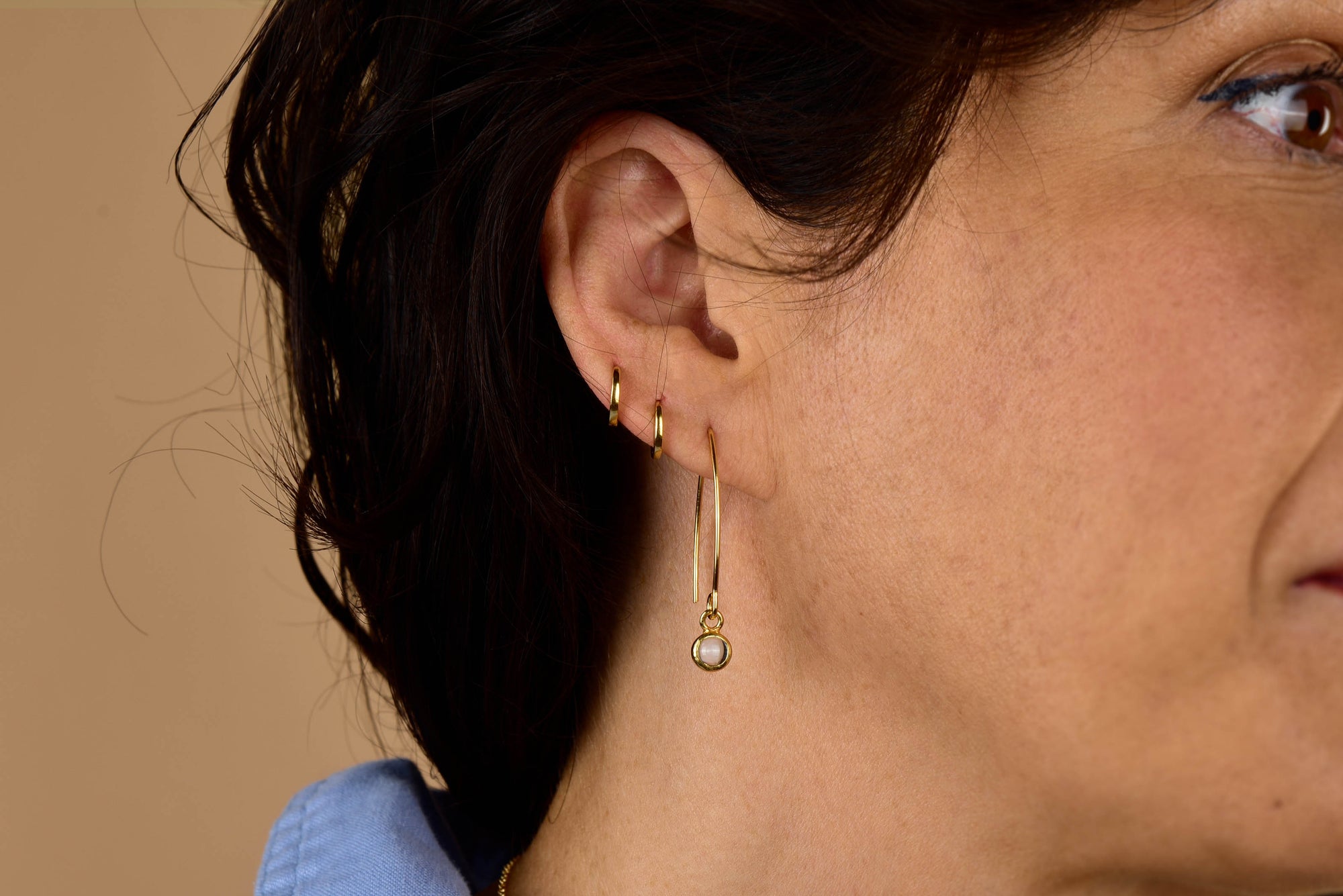 Close-up of a woman's ear adorned with three gold hoop earrings, the Rose Quartz Gem Dot Arc Earrings featuring a small round rose quartz pendant, and short dark hair. She is wearing a light blue top.