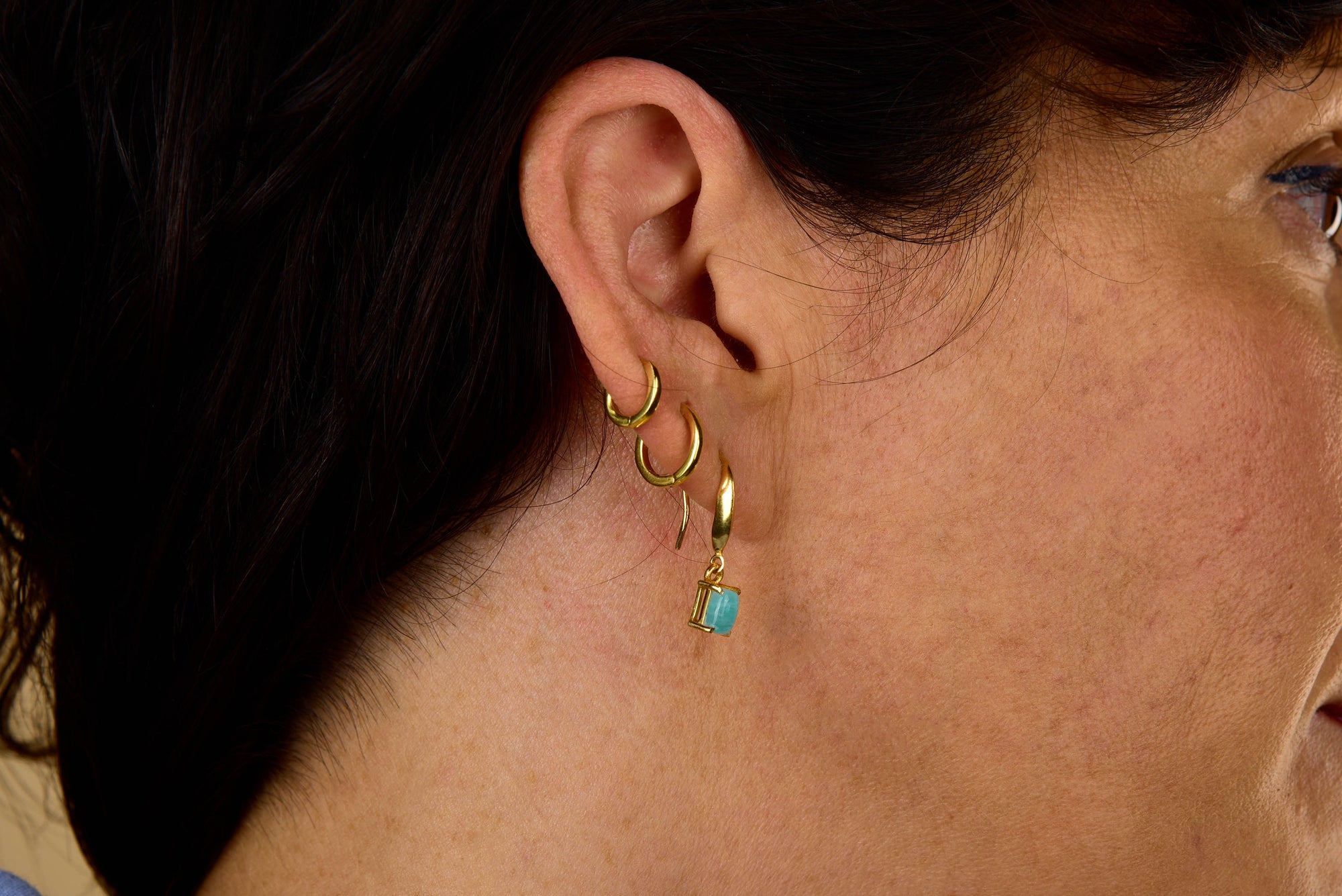 Close-up of a woman's ear with three gold hoop earrings and one Amazonite Octavia Drop Earring—featuring an elegant octagonal amazonite gemstone—set against her brown hair and light skin.