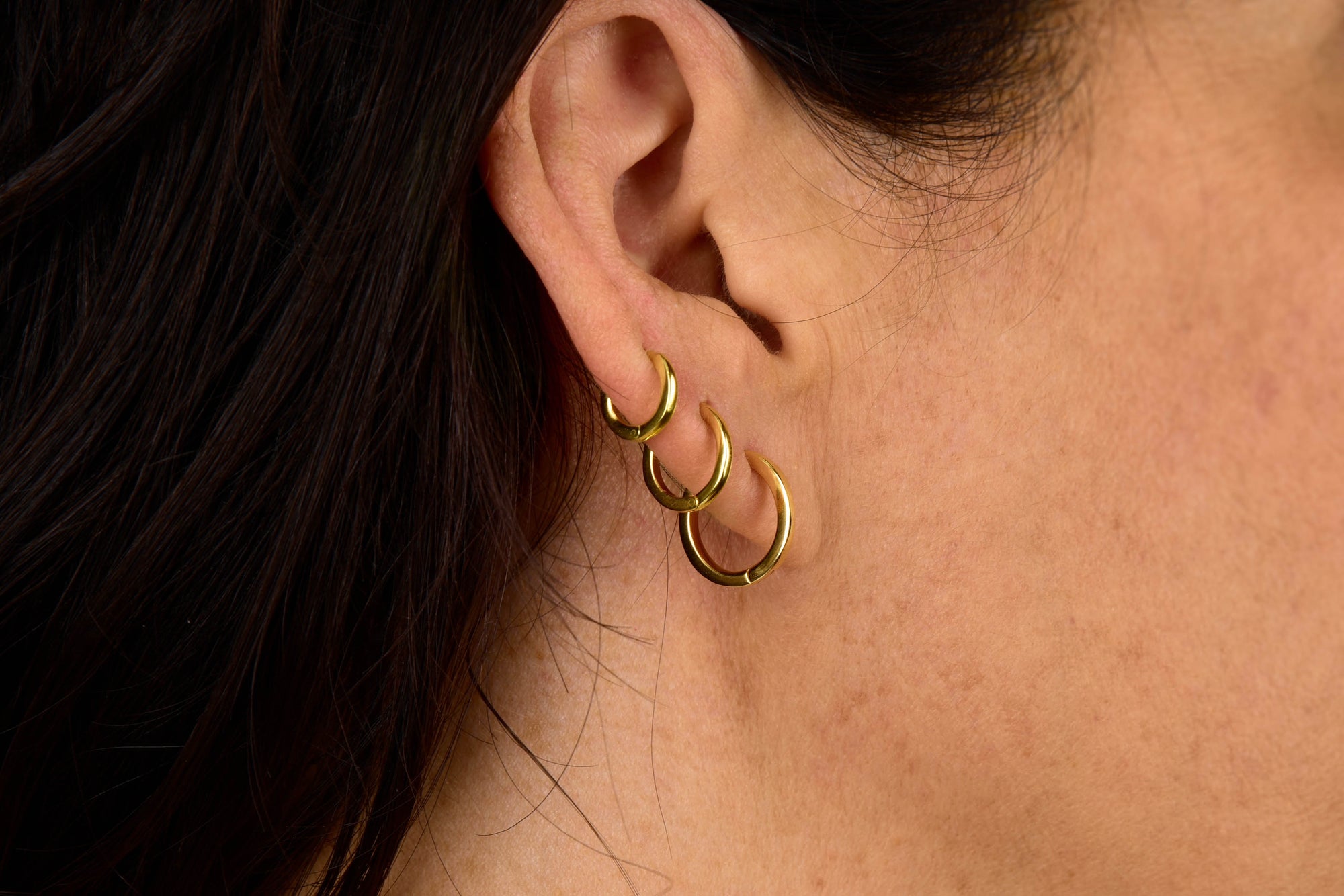 Close-up of a person’s ear adorned with three Nell Huggie Earrings in gold-plated silver. The individual has dark hair and light skin, and the minimalist hoops are arranged vertically along the outer ear.