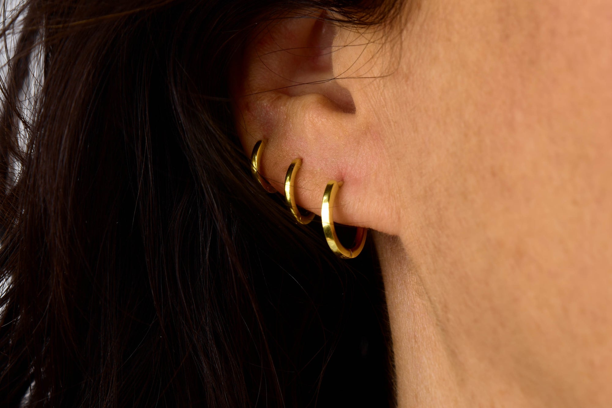 Close-up of a person’s ear adorned with four small gold hoop earrings, including the minimalist Nell Huggie Earrings, lined up along the outer edge. The person’s dark hair partially covers their ear.