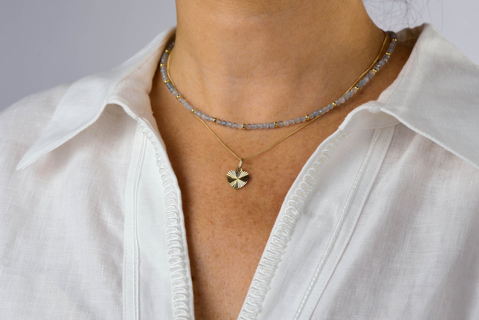 A person wearing a white linen shirt with two layered necklaces: one featuring small gray beads and a gold-plated sunburst pendant, complemented by the Celeste Heart Necklace.