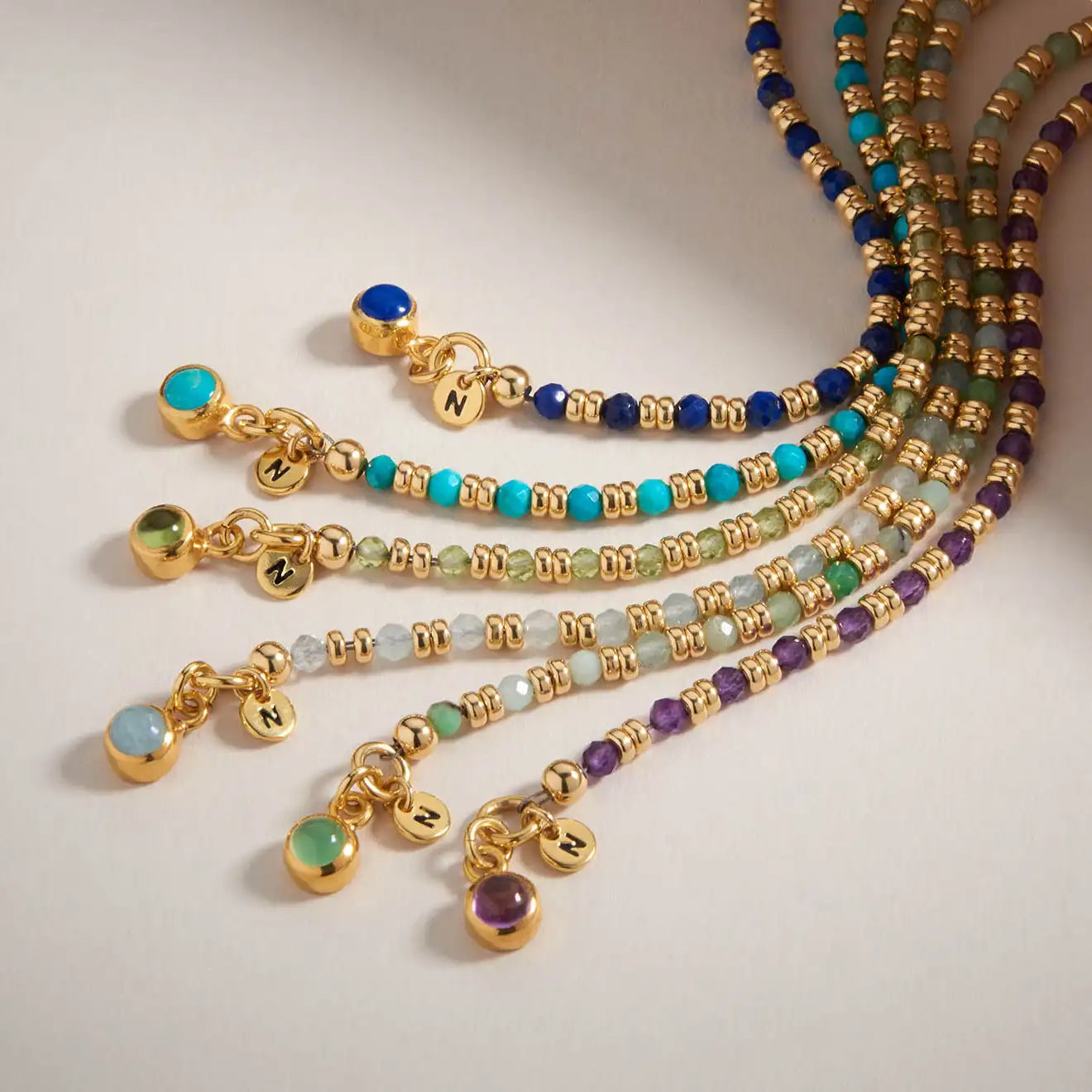 Seven beaded gold bracelets with different colored stones and gold charms, each engraved with the letter “N,” arranged in a fan shape on a neutral background.