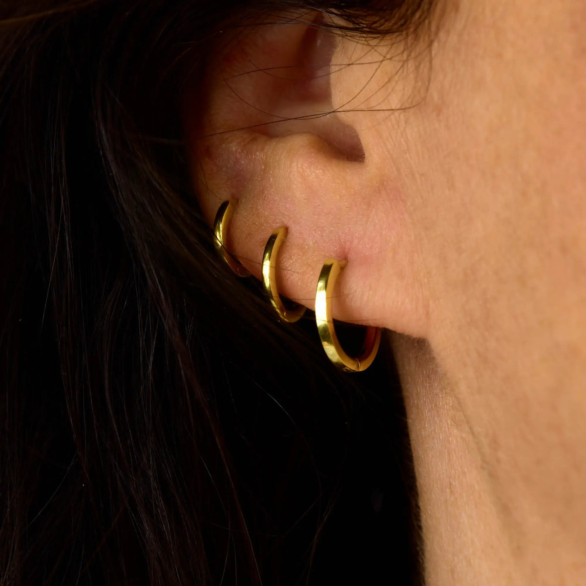 Close-up of an ear with three gold hoop earrings in the lobe and upper cartilage. The persons hair is dark and partially covers the ear.