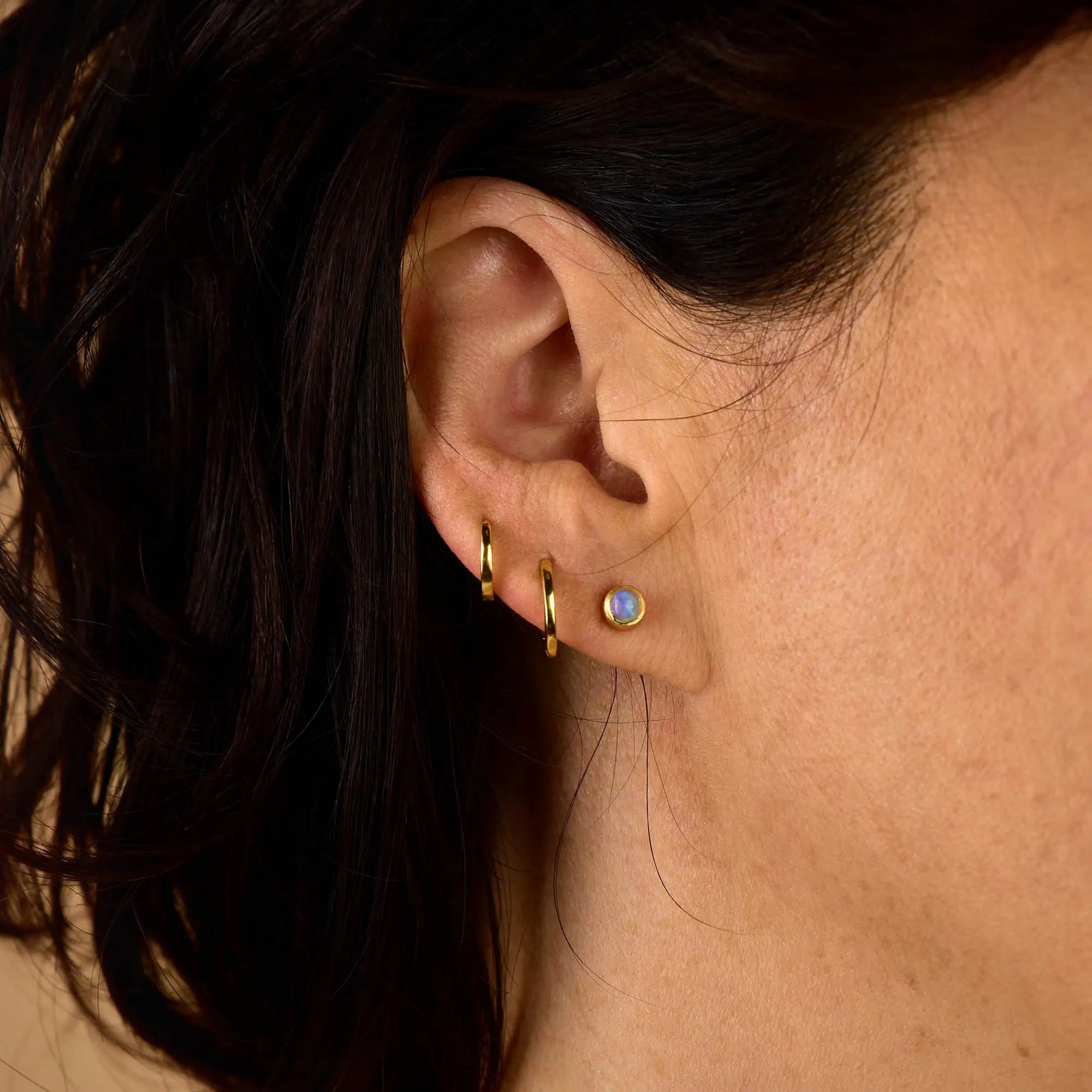 Close-up of a woman’s ear adorned with three small gold hoops and one Mini Gem Dot Birthstone Earring featuring a round opal stone in sterling silver. Her dark hair partially covers the ear, while the softly blurred background highlights the subtle elegance of this birthstone earring.