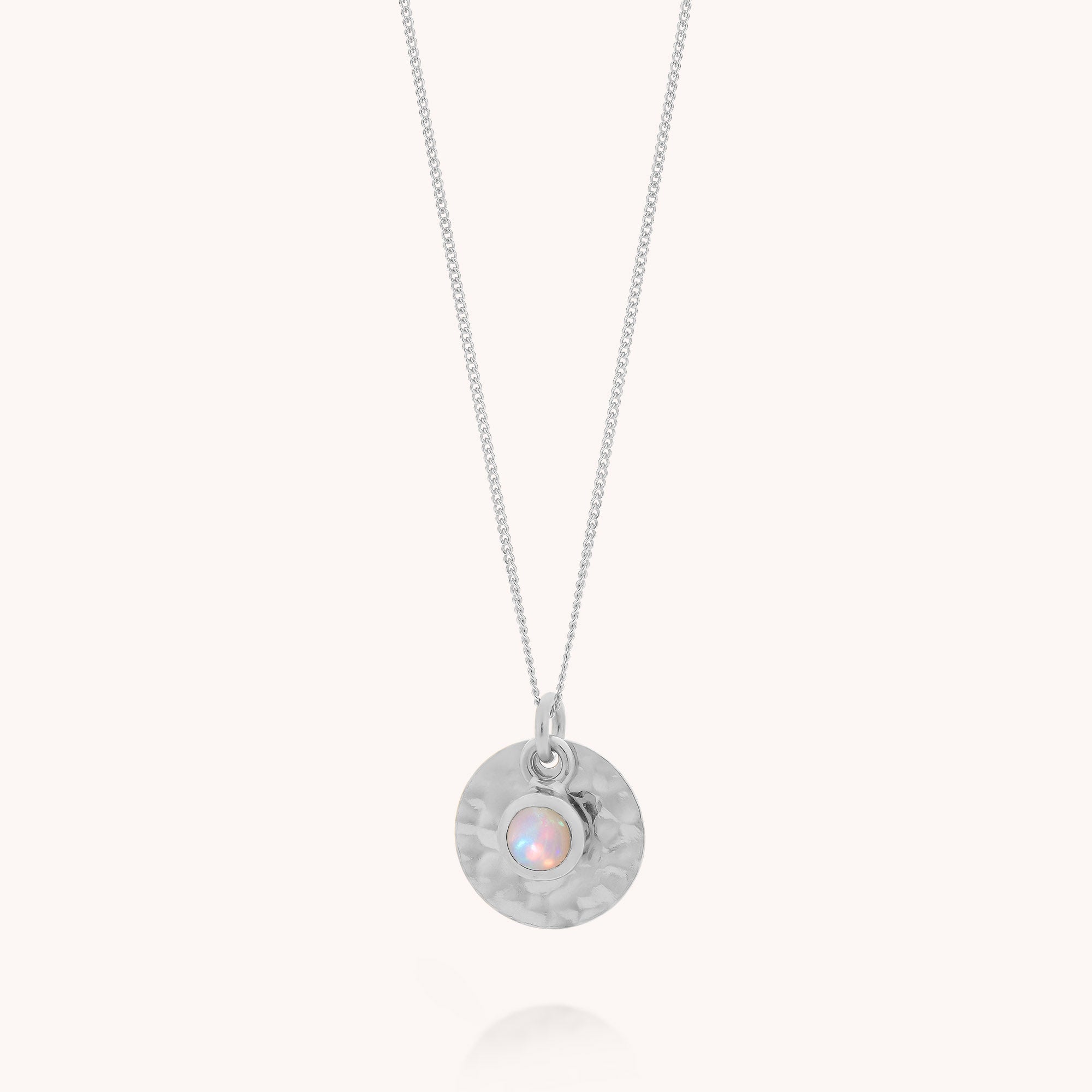 The Ethiopian Opal Luna Disc Necklace is elegantly displayed against a plain, light background, showcasing its round, hammered silver pendant. At the center shines a delicate Ethiopian opal—the October birthstone—infusing the piece with iridescent beauty and timeless significance.