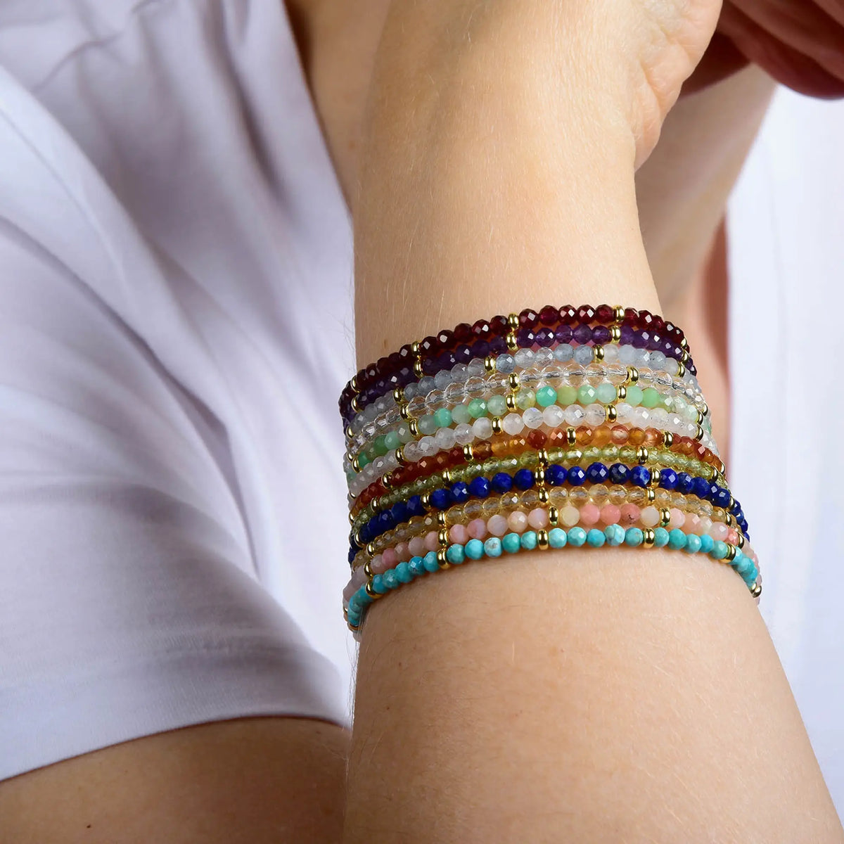 A person wearing a white shirt displays their forearm adorned with multiple colorful beaded bracelets in various shades and patterns, including the striking Garnet Orbit Bracelet - January, which highlights the rich red hue of the January birthstone alongside touches of sterling silver accents.