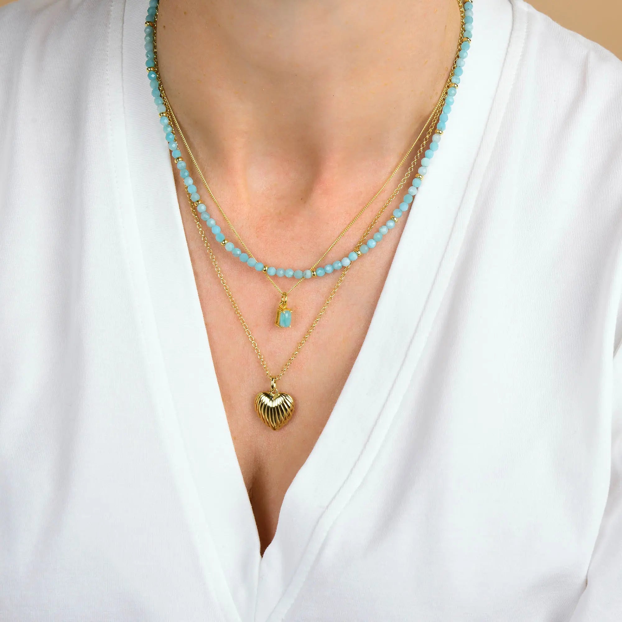 A person wearing a white V-neck top and three layered necklaces: one is the Scallop Heart Necklace featuring a gold heart pendant, another is adorned with blue beads and gold chains, and the third is a sterling silver necklace with a small blue rectangular charm.