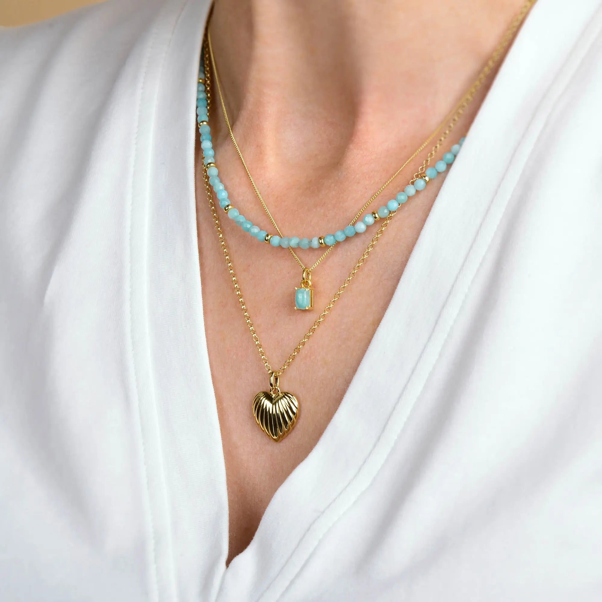 A person wearing a white V-neck shirt and three layered necklaces: a blue beaded strand, a gold chain with a small blue pendant, and a gold chain featuring the Scallop Heart Necklace with its textured scalloped heart pendant.