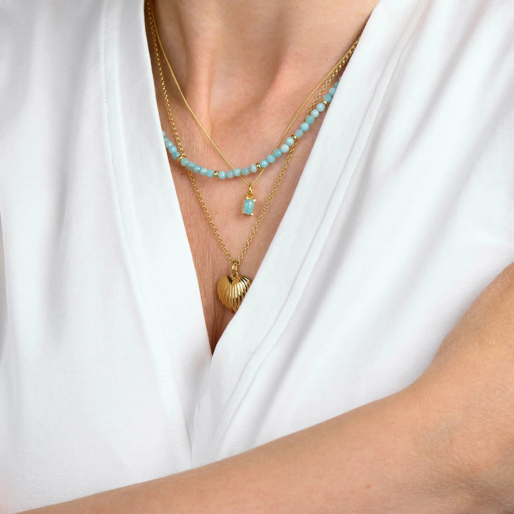 A person wearing a white V-neck top and three gold necklaces: one with blue beads, one featuring the Amazonite Octavia Necklace with a blue stone pendant, and one with a gold shell pendant.