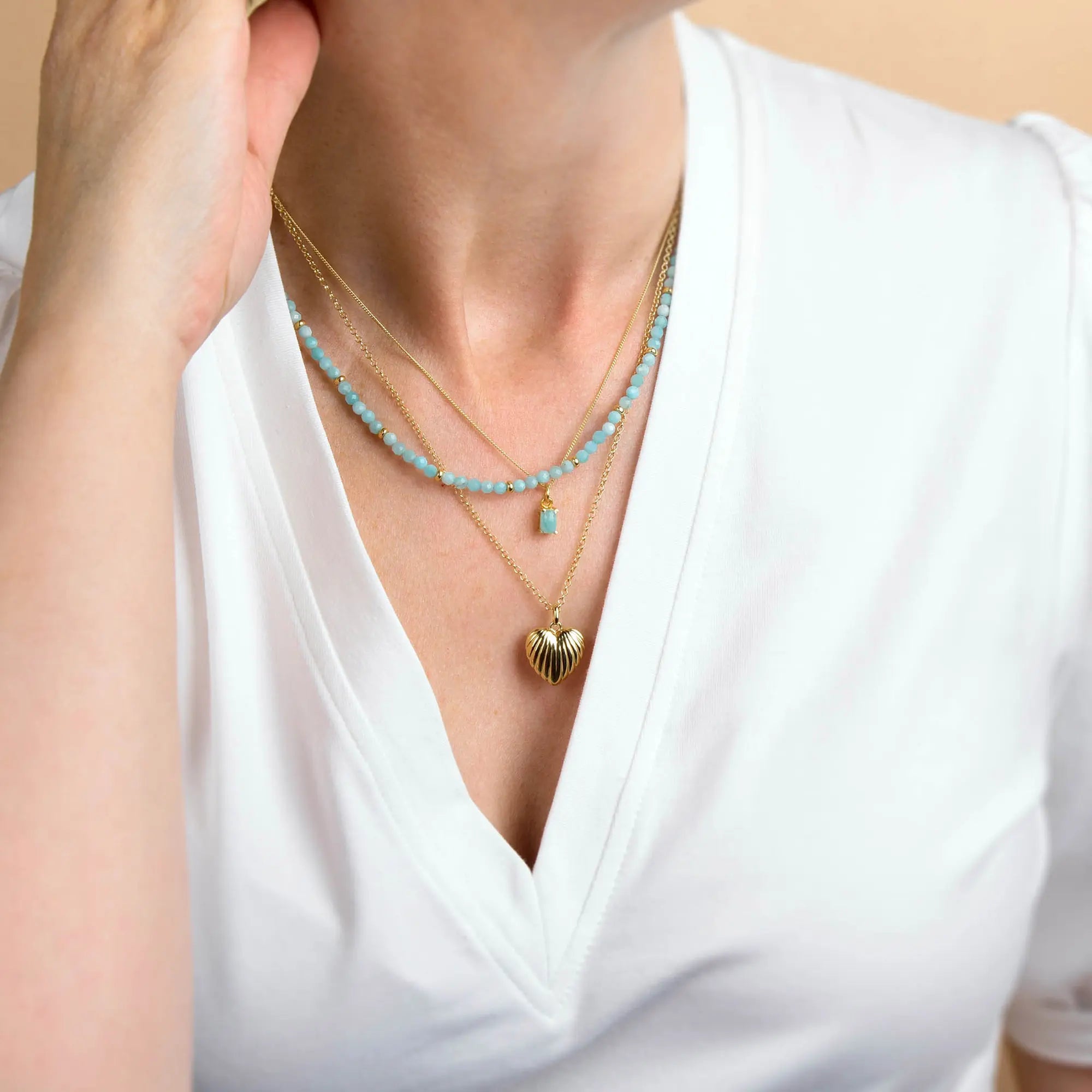 Wearing a white V-neck shirt, the individual showcases three layered necklaces: one accented with blue natural gemstone beads, one featuring the Amazonite Orbit Necklace as a delicate pendant, and another with a gold heart-shaped charm—highlighting the elegance of handcrafted jewellery.