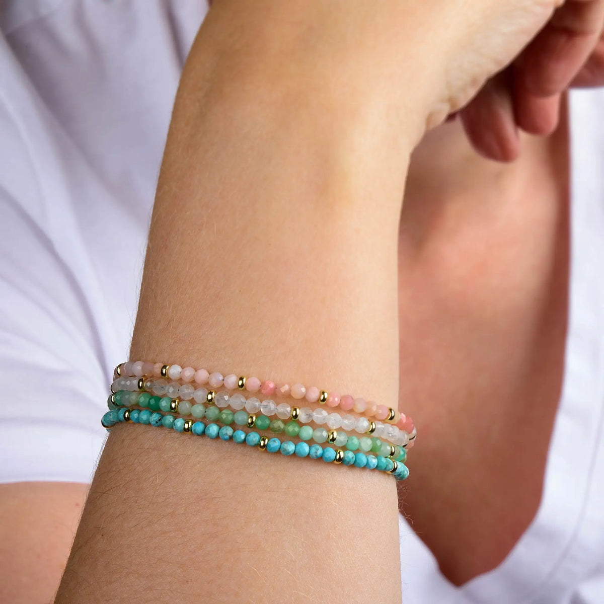 A person wearing a white shirt rests their chin on their hand, displaying three beaded bracelets—including the vibrant green Chrysoprase Orbit Bracelet - May, the May birthstone—alongside pink and turquoise designs accented with small gold details; all pieces are part of a stunning collection of handcrafted gemstone jewelry.