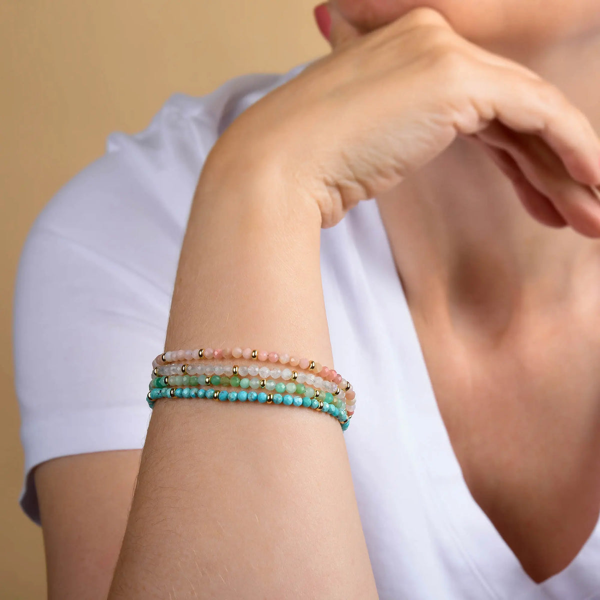 A person wearing a white shirt rests their chin on their hand, displaying four colorful handcrafted bracelets on their wrist, including the Rainbow Moonstone Orbit Bracelet - June, which glimmers softly in the light.