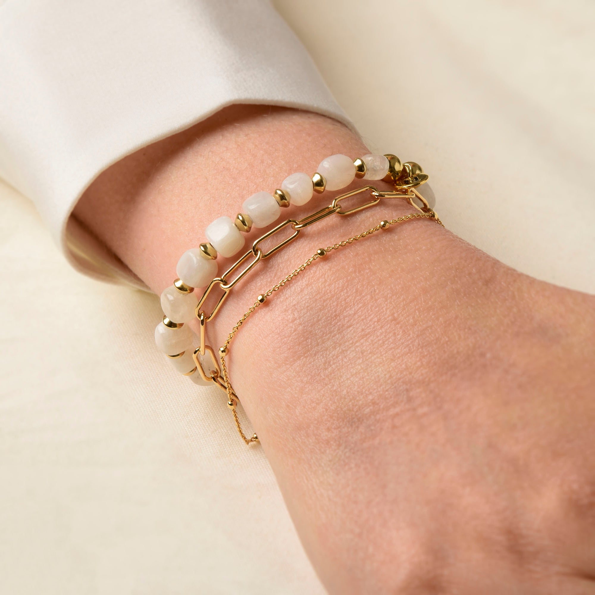 A close-up of a wrist wearing three bracelets: a white beaded bracelet, a gold chain-link bracelet, and the Satellite Bracelet—a delicate gold plated sterling silver chain adorned with small beads. The person is dressed in a light-colored long sleeve.