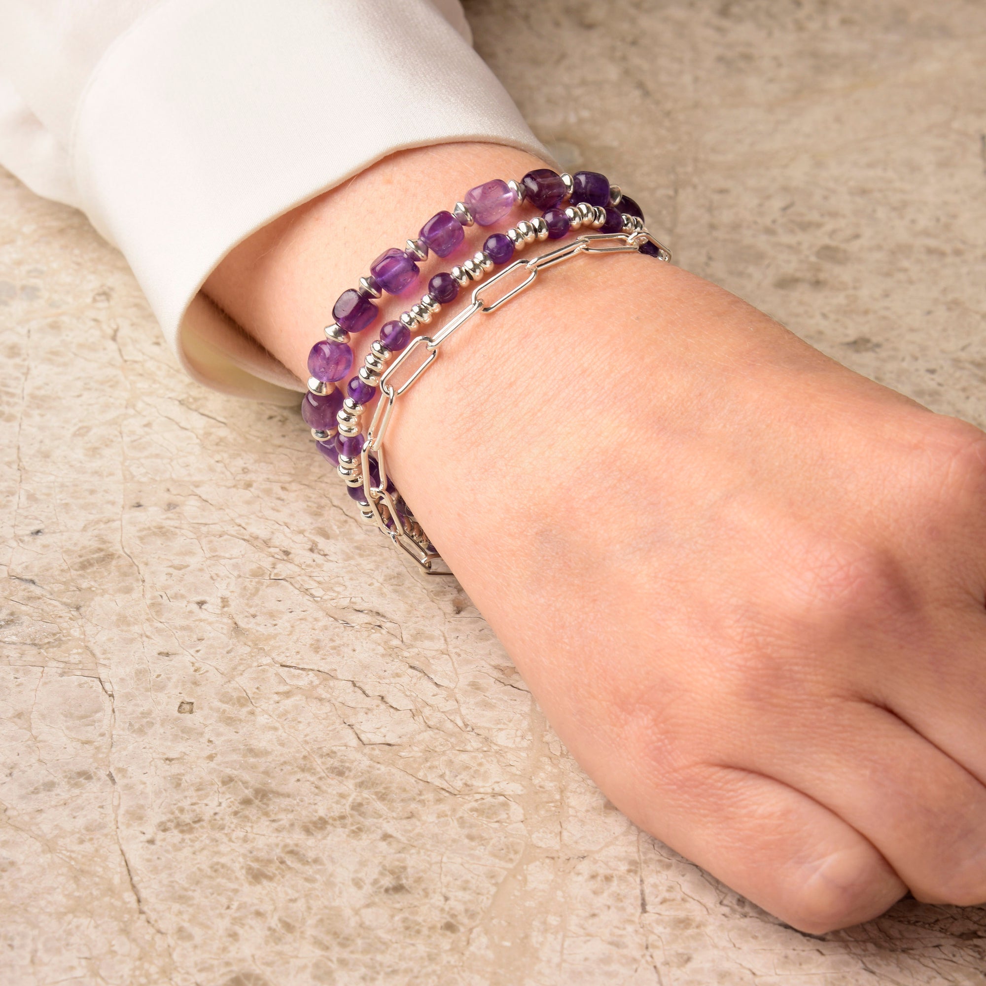 A hand wearing a white long sleeve shirt rests on a marble surface, adorned with several Gem Birthstone Bracelets stacked on the wrist, featuring natural gemstone beads in silver and purple tones with elegant gold plated sterling silver accents.