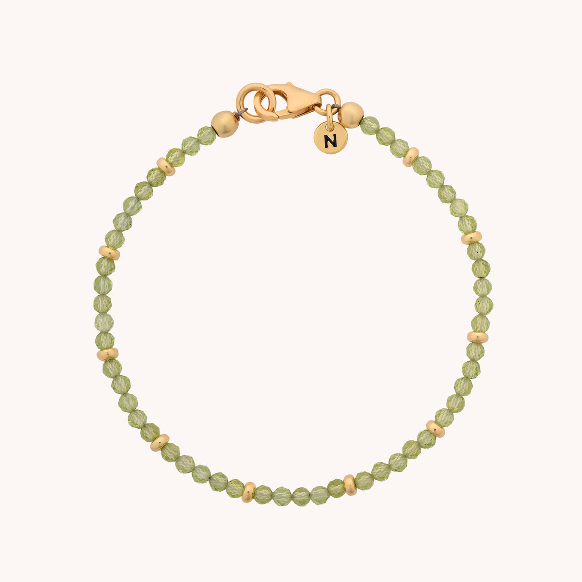 Introducing the Peridot Orbit Bracelet - August: a delicate piece adorned with small green peridot beads and elegant gold accents. It features a gold lobster clasp and is finished with a round charm engraved with the letter N, beautifully highlighting the vibrant hue of the August birthstone.