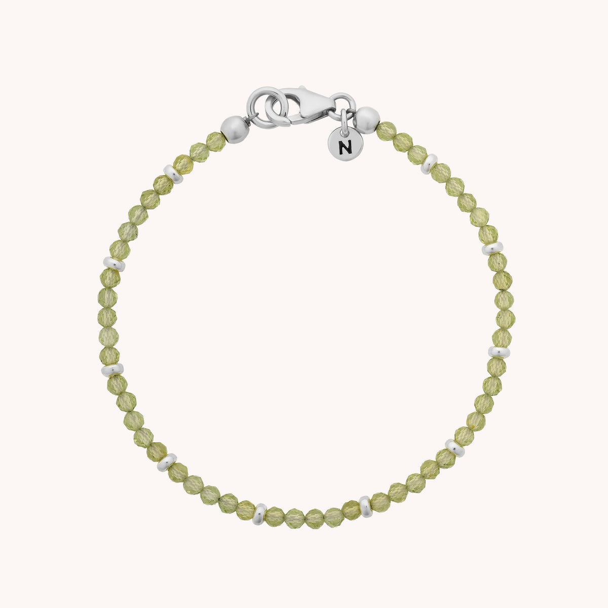 The Peridot Orbit Bracelet - August showcases delicate light green peridot beads—evoking the beauty of the August birthstone—adorned with petite silver accents, a silver lobster clasp, and a round charm engraved with the letter N, all displayed in a circular arrangement on a plain background.