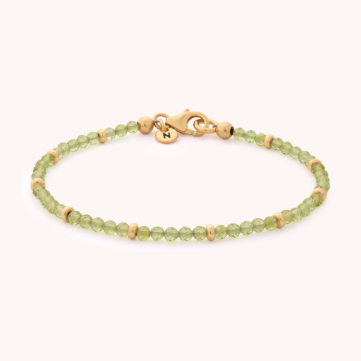 The Peridot Orbit Bracelet - August features delicate green beads paired with small gold spacer beads and a gold clasp, highlighted by a round gold charm engraved with the letter N. Inspired by the vibrant hue of the August birthstone, this elegant bracelet is shown against a simple, light-colored background.