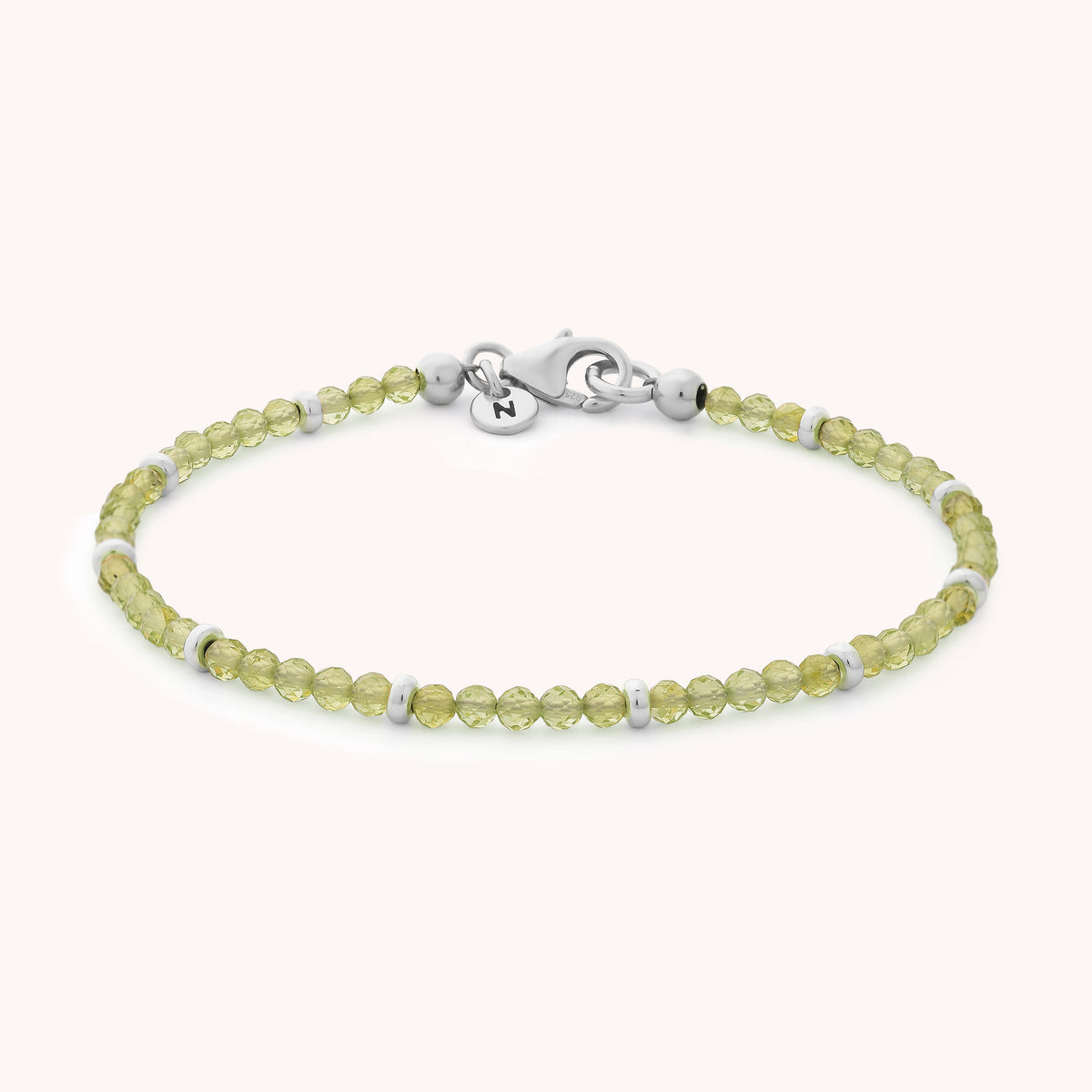 The Peridot Orbit Bracelet - August showcases small, round light green peridot beads—the August birthstone—accented with petite silver rings and finished with a sleek silver clasp and a discreet tag. This delicate bracelet is elegantly presented against a plain light background.