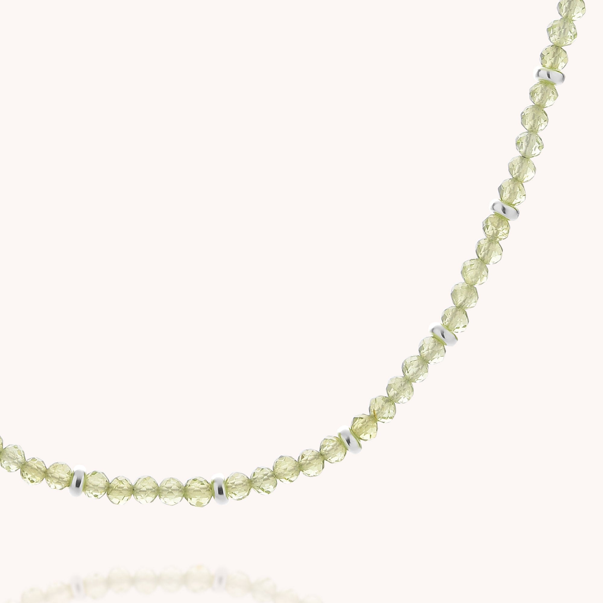 A close-up of the Peridot August Birthstone Orbit Necklace, featuring delicate light green faceted peridot beads accented with small silver spacers evenly arranged throughout, displayed on a plain white background. This elegant necklace beautifully showcases the natural sparkle and charm of the August birthstone.