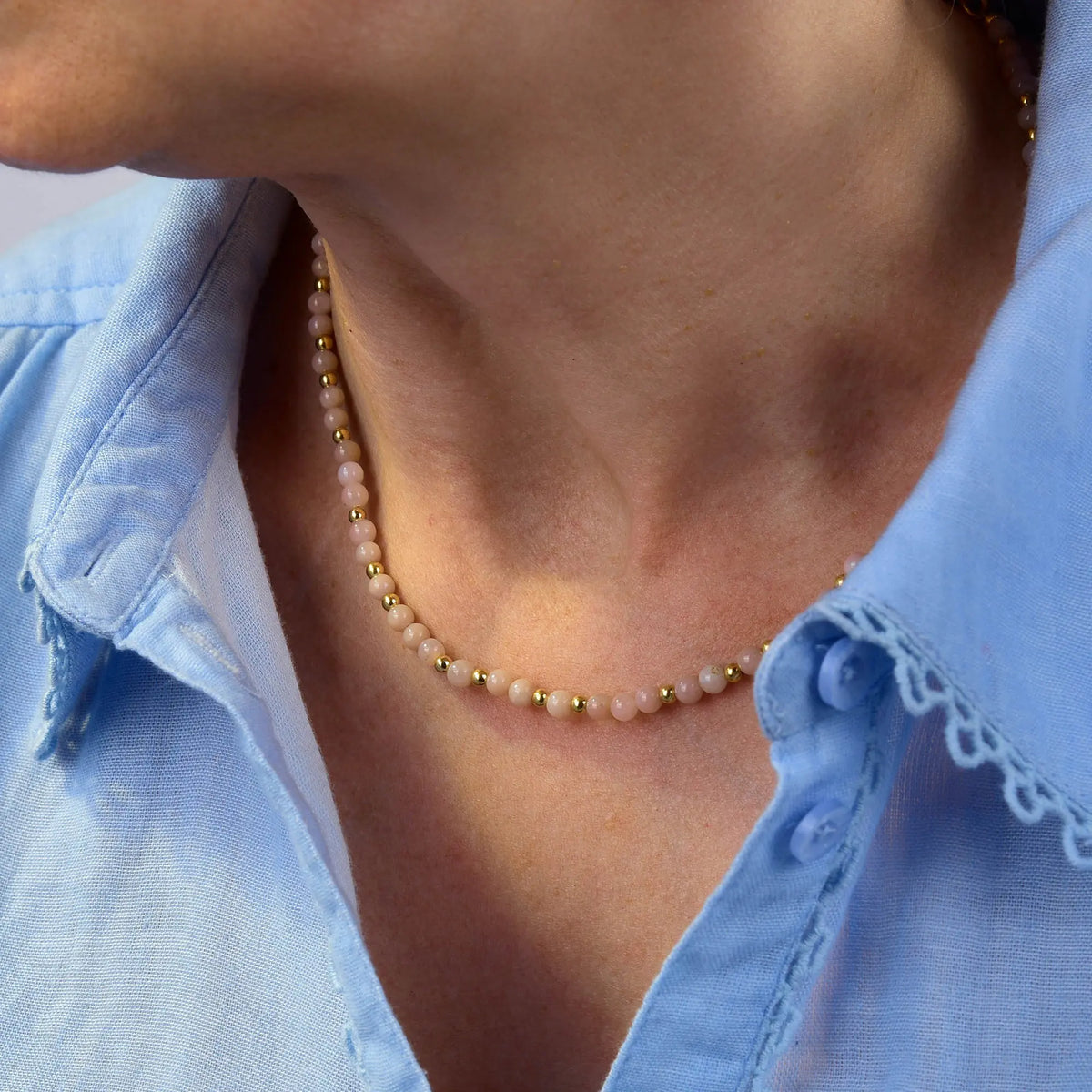 A person wearing a light blue collared shirt and a delicate handcrafted beaded necklace—featuring the Pink Opal October Birthstone Orbit Necklace with small gold and pale pink beads—around their neck.