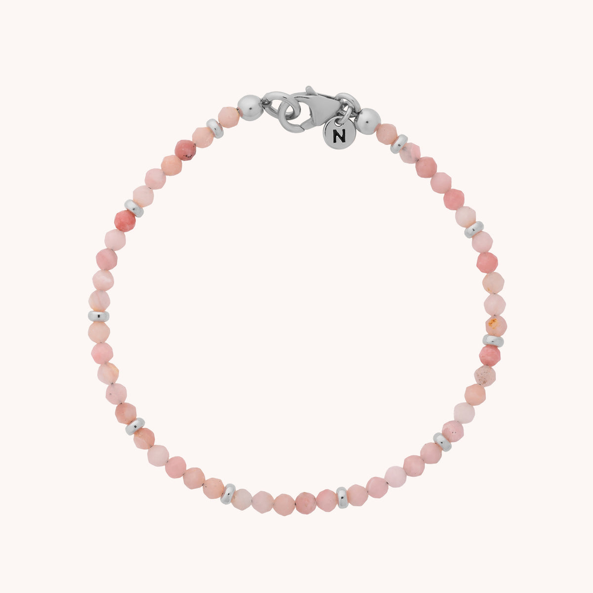 The Pink Opal Orbit Bracelet - October is a delicate, handcrafted piece featuring small pink opal beads reminiscent of the October birthstone, spaced by tiny silver beads. It is finished with a silver clasp adorned with a round charm engraved with the letter N and is shown laid flat against a light background.