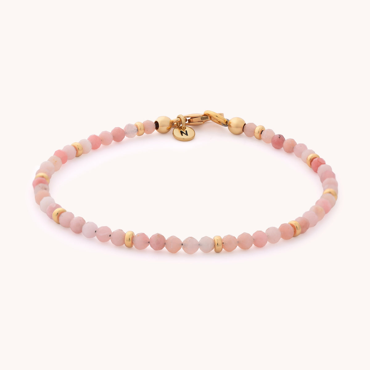 The Pink Opal Orbit Bracelet - October is a delicate, handcrafted piece featuring small pink opal beads, gold accents, and a gold clasp adorned with a round charm engraved with the letter N on a light background—making it a perfect accessory for those with an October birthday.