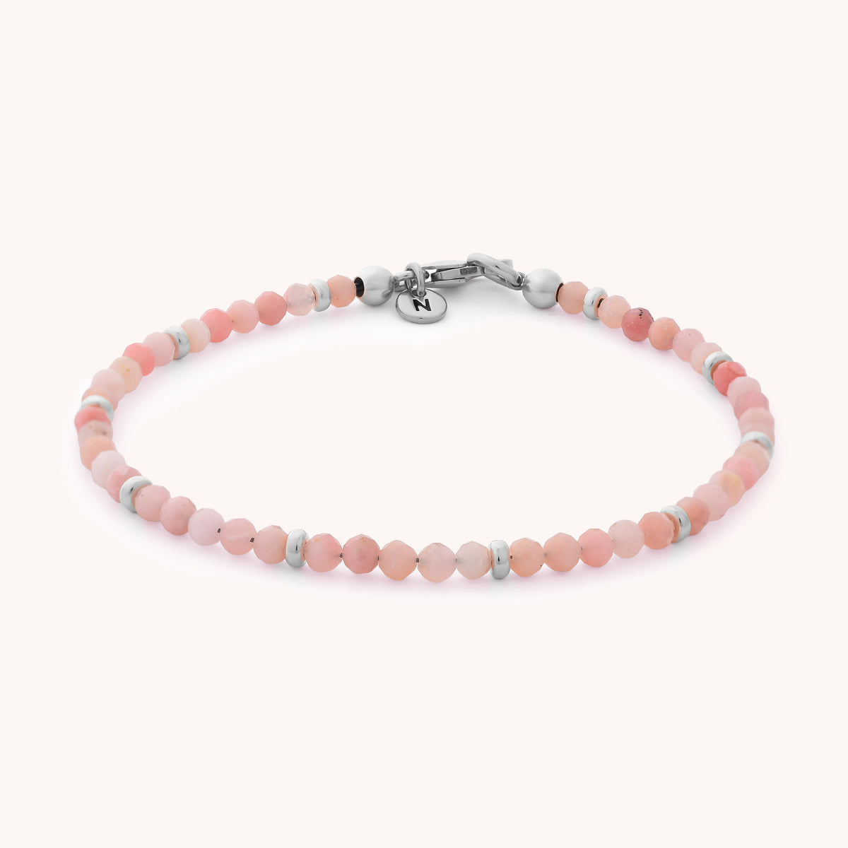 The Pink Opal Orbit Bracelet - October features small, round pink opal beads accented with silver spacers and a secure metal clasp. A dainty round charm engraved with the letter “N” hangs near the clasp, making this bracelet a thoughtful October birthstone gift. Set against a light, neutral background, its handcrafted elegance shines through.
