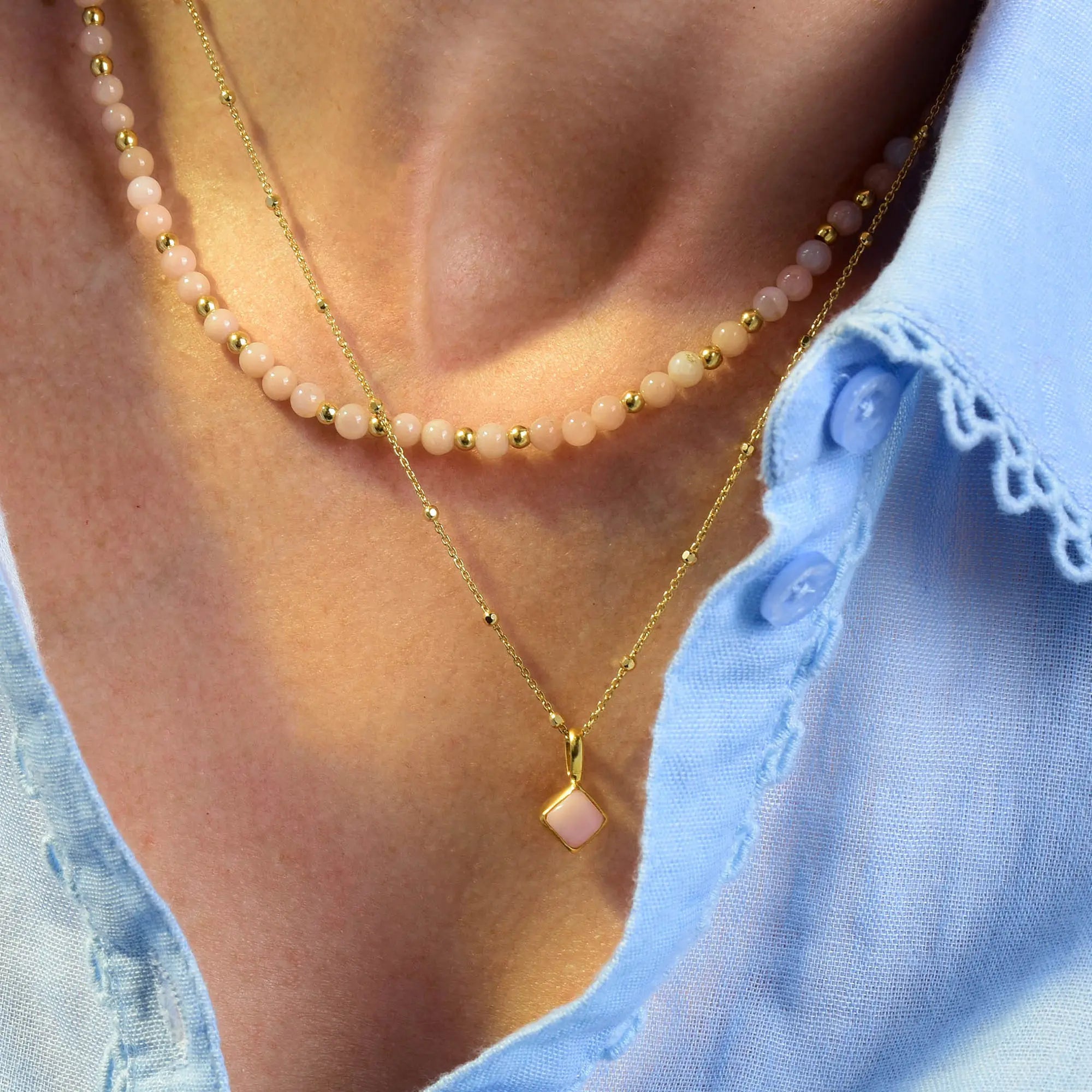 A close-up of a person wearing a blue collared shirt and two necklaces: one is the Pink Opal Diamond Birthstone Necklace October, featuring delicate pink opal stones with gold accents and pink beads, while the other is a gold chain with a square pink pendant.