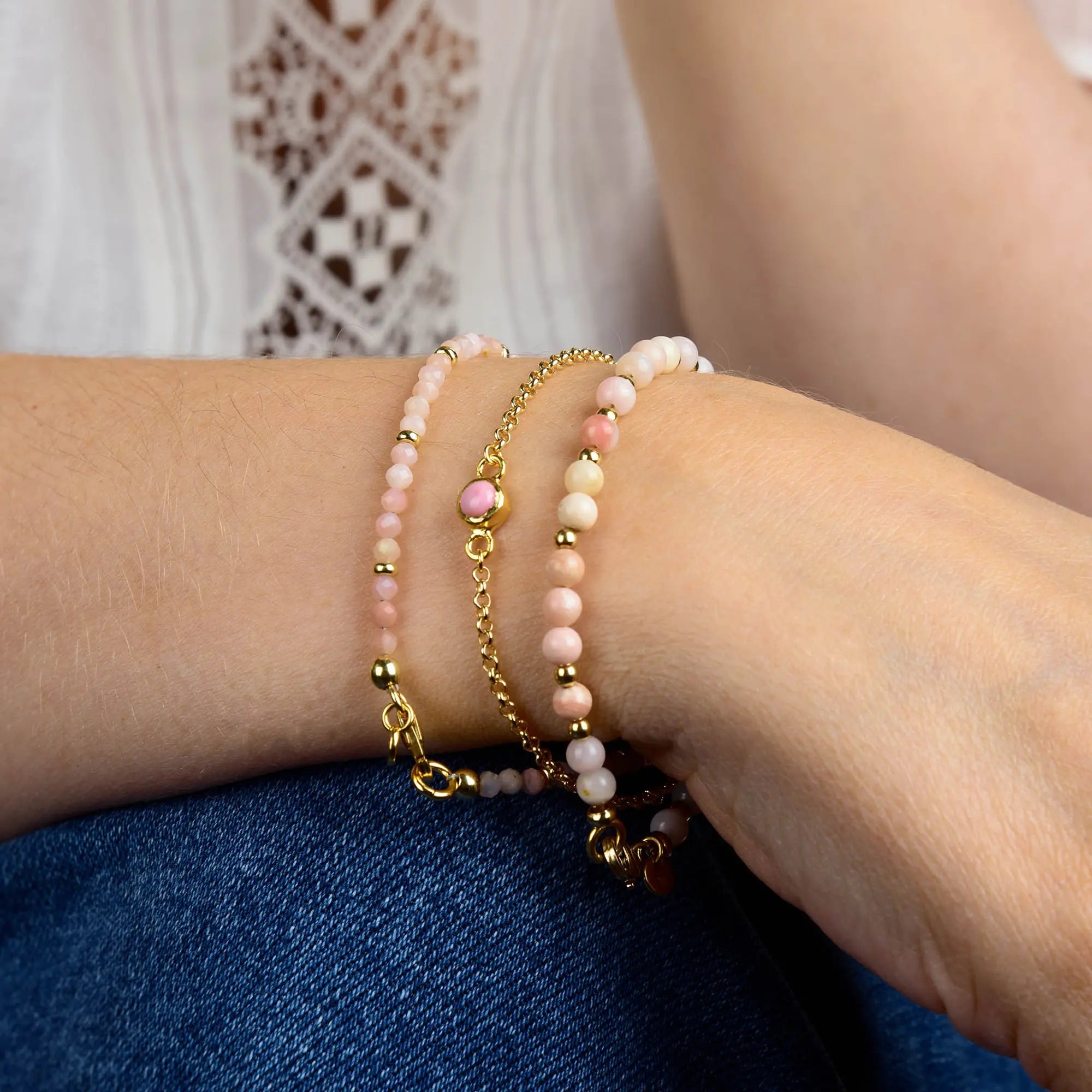 A person wearing three delicate beaded bracelets—including the Pink Opal Birthstone Gem Dot Bracelet - October—in soft pink, white, and gold tones rests their arm on their leg, with a white lace blouse and blue jeans visible.