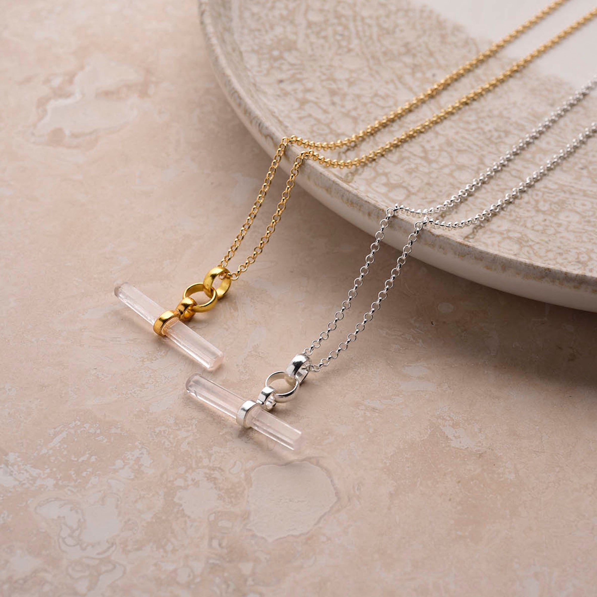 Two Gemstone T-Bar Necklace - Quartz pieces, one on a gold chain and the other on sterling silver, feature horizontal clear quartz pendants and are displayed on a textured beige surface near a ceramic plate, highlighting elegant jewellery.