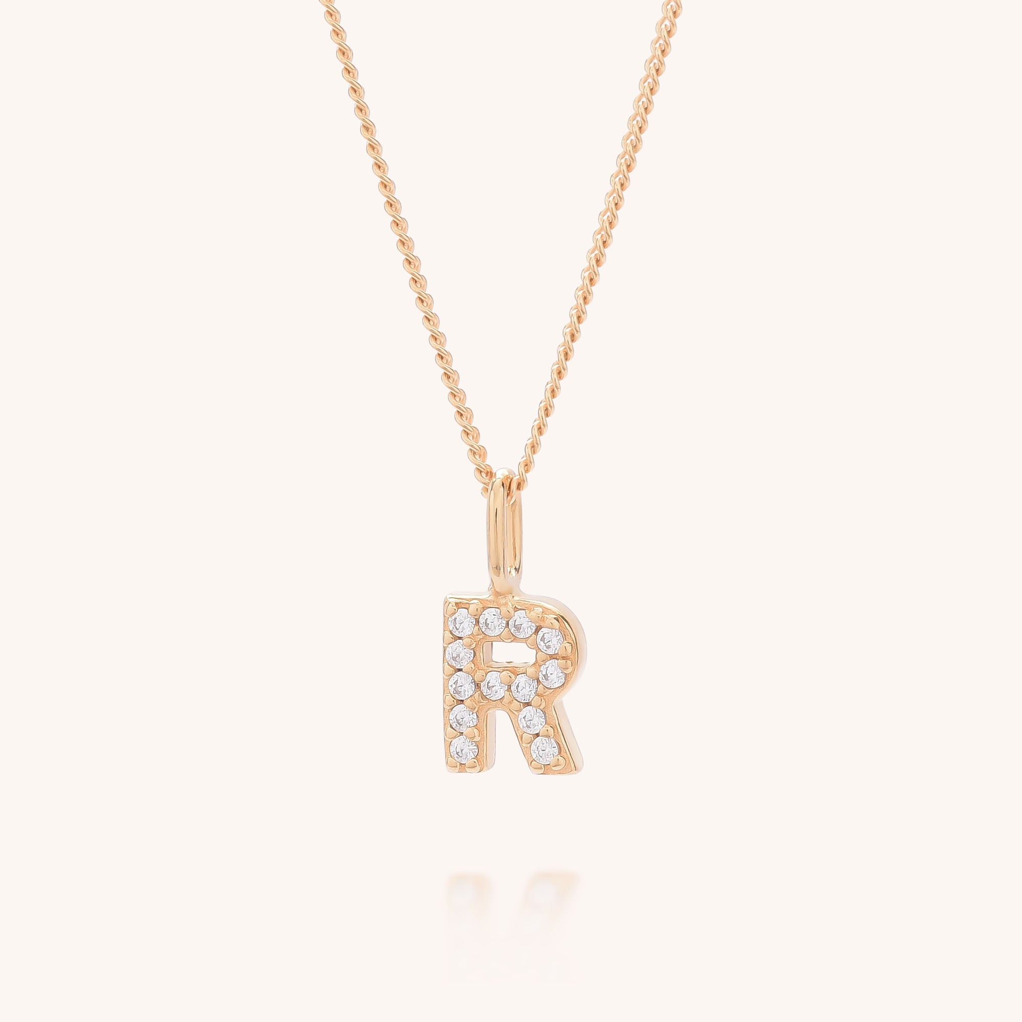 The Initial Sparkle Necklace features a gold letter R pendant adorned with sparkling cubic zirconia stones, shown on a light beige background.