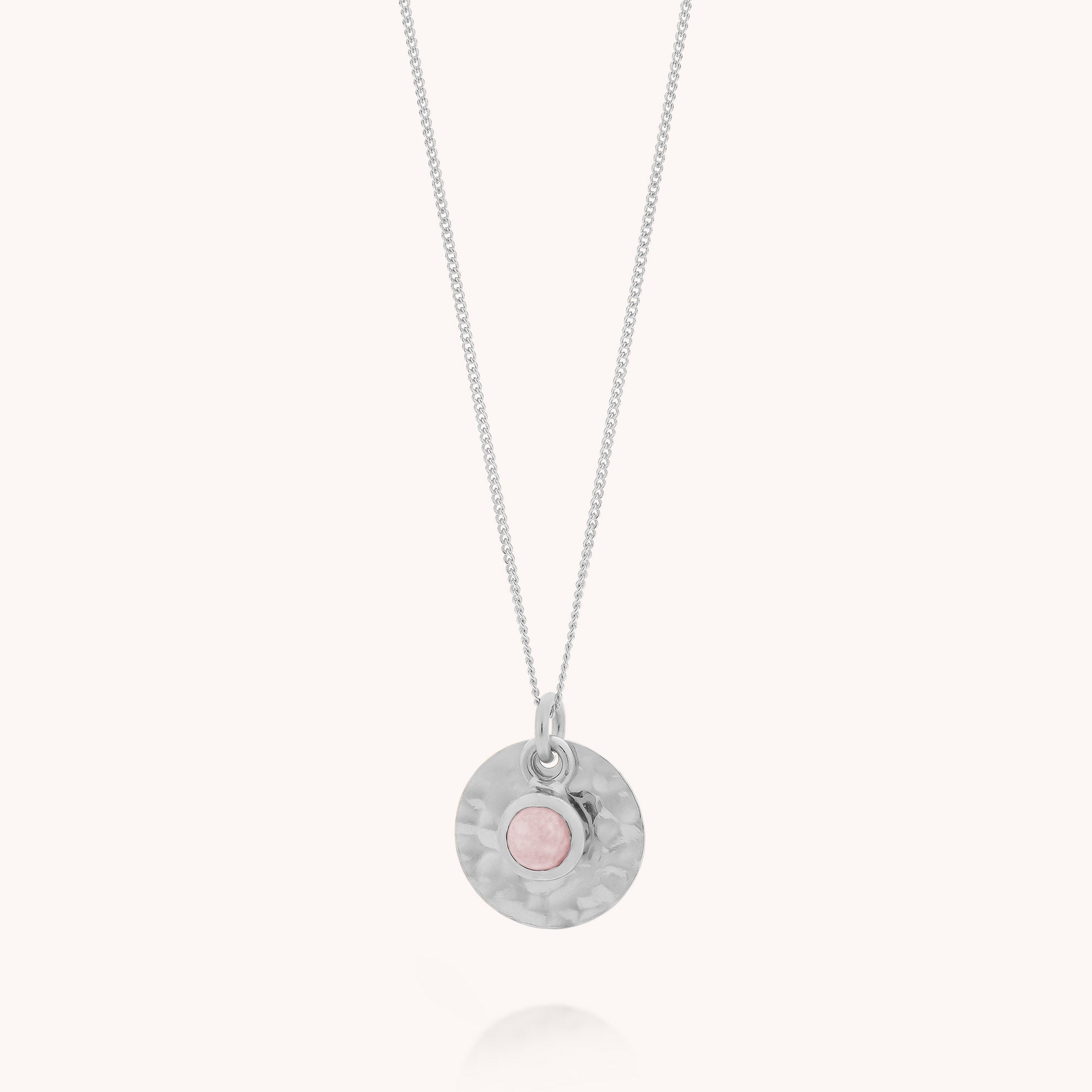 The Rose Quartz Luna Disc Necklace showcases a silver hammered round pendant with a small pink stone set in the center, presented against a simple light background.