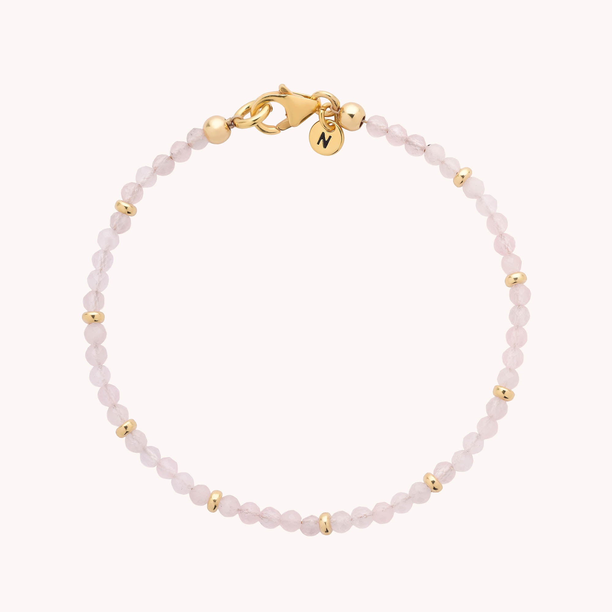 The Rose Quartz Orbit Bracelet features delicate, small faceted rose quartz beads accented by gold spacers, a gold clasp, and a small round gold charm engraved with the letter N.