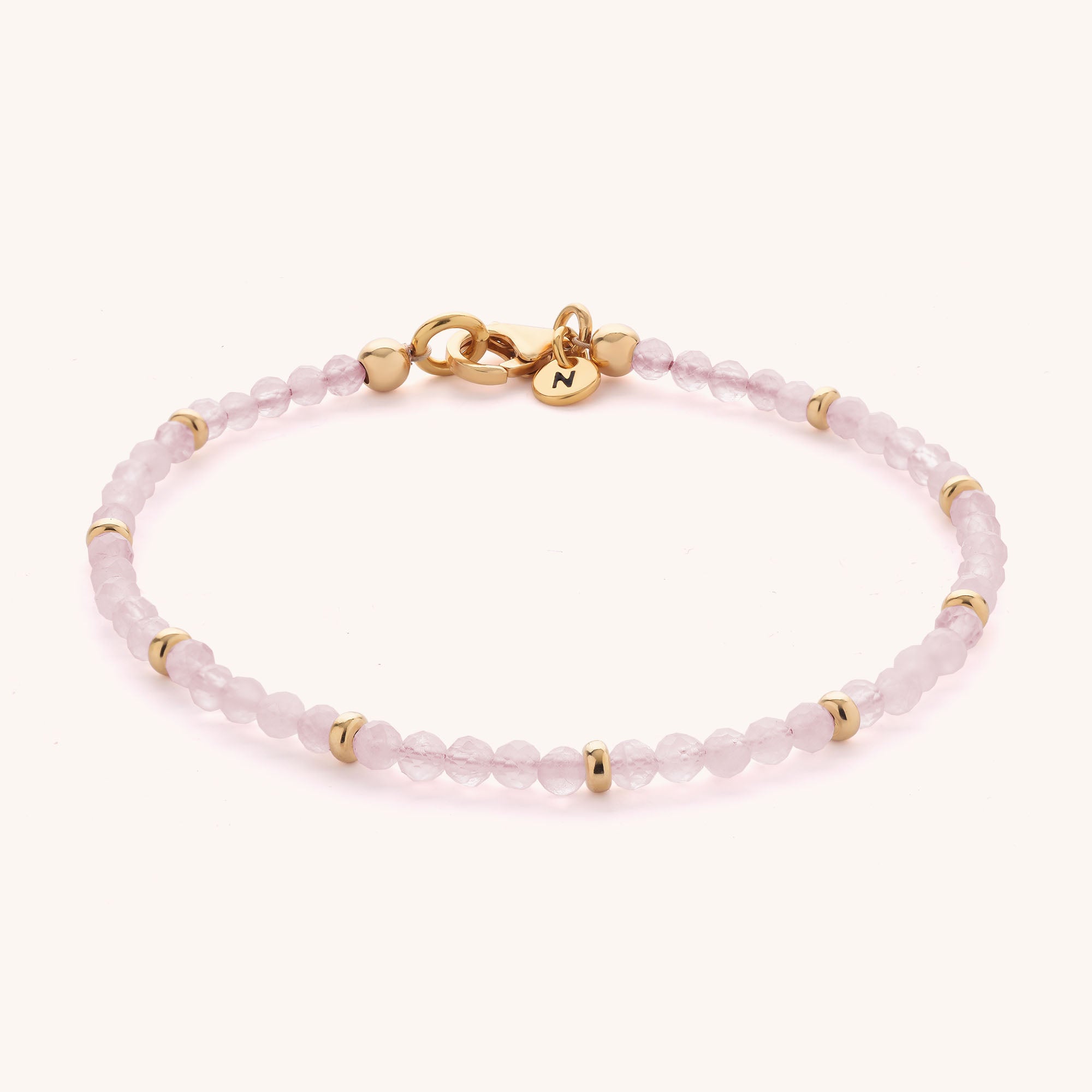 The Rose Quartz Orbit Bracelet is crafted with light pink, faceted rose quartz beads accented by small gold rings. It features a gold clasp and a round gold charm engraved with the letter N.