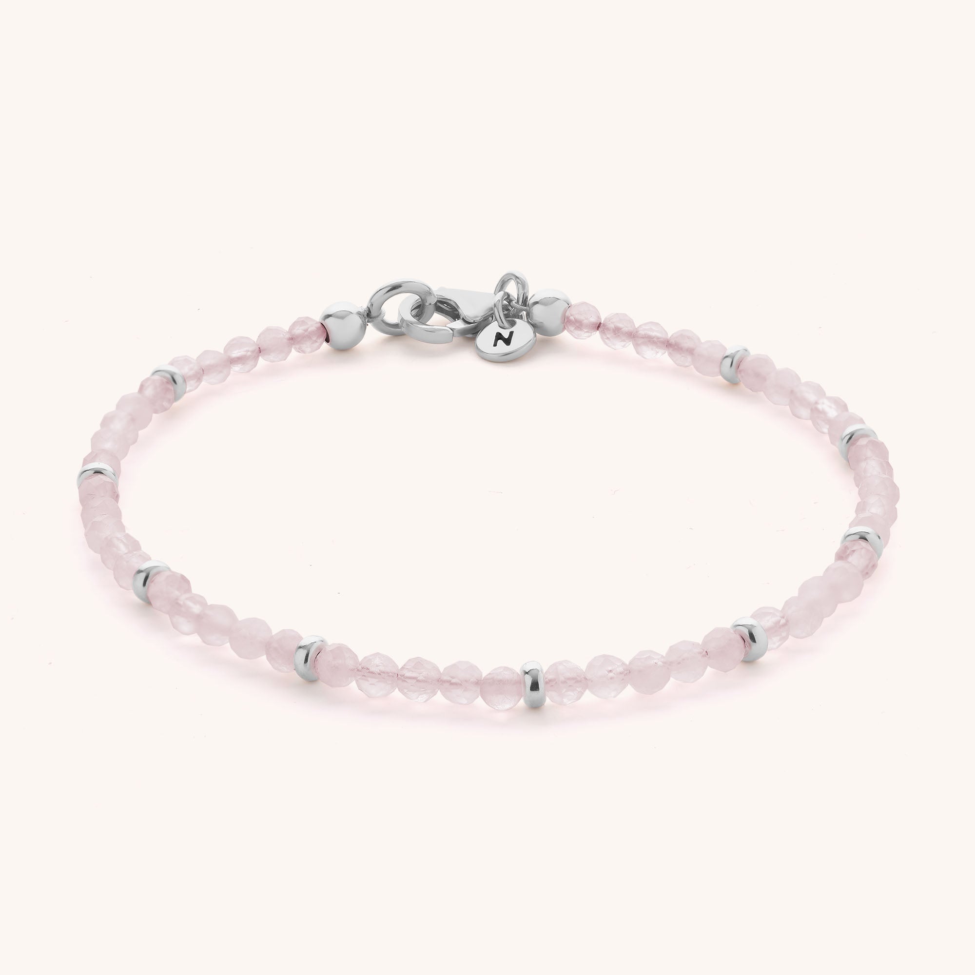 The Rose Quartz Orbit Bracelet features small, round, pale pink faceted rose quartz beads accented with sterling silver spacers and finished with a classic silver clasp. A small round charm engraved with the letter N hangs near the clasp, all set against a light off-white background.