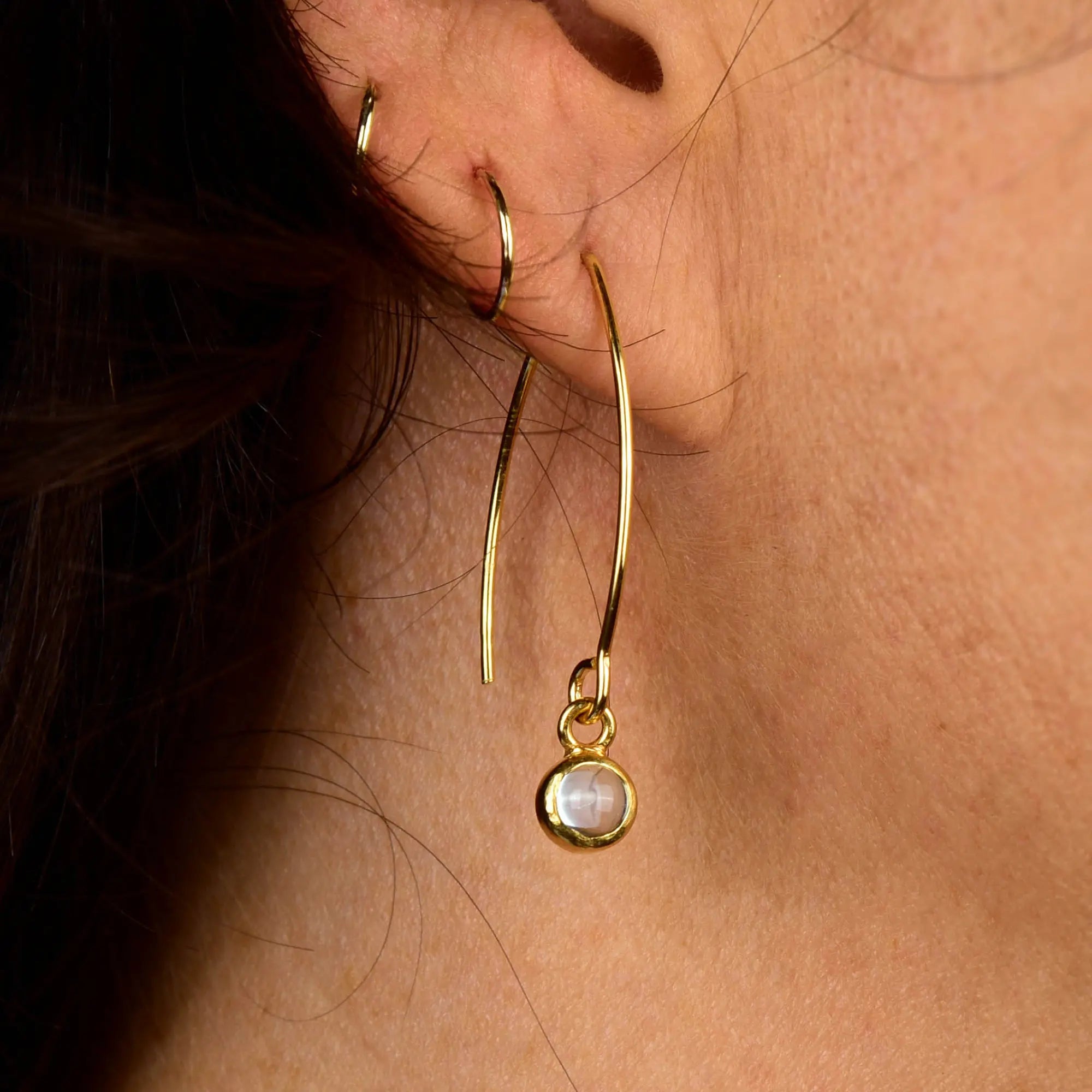 Close-up of a person’s ear adorned with three gold earrings: two thin gold hoops and one striking Rose Quartz Gem Dot Arc Earring, featuring a dangling rose quartz gemstone set in a gold frame. The background reveals part of the person’s hair and skin, providing an elegant setting for these unique gemstone earrings.