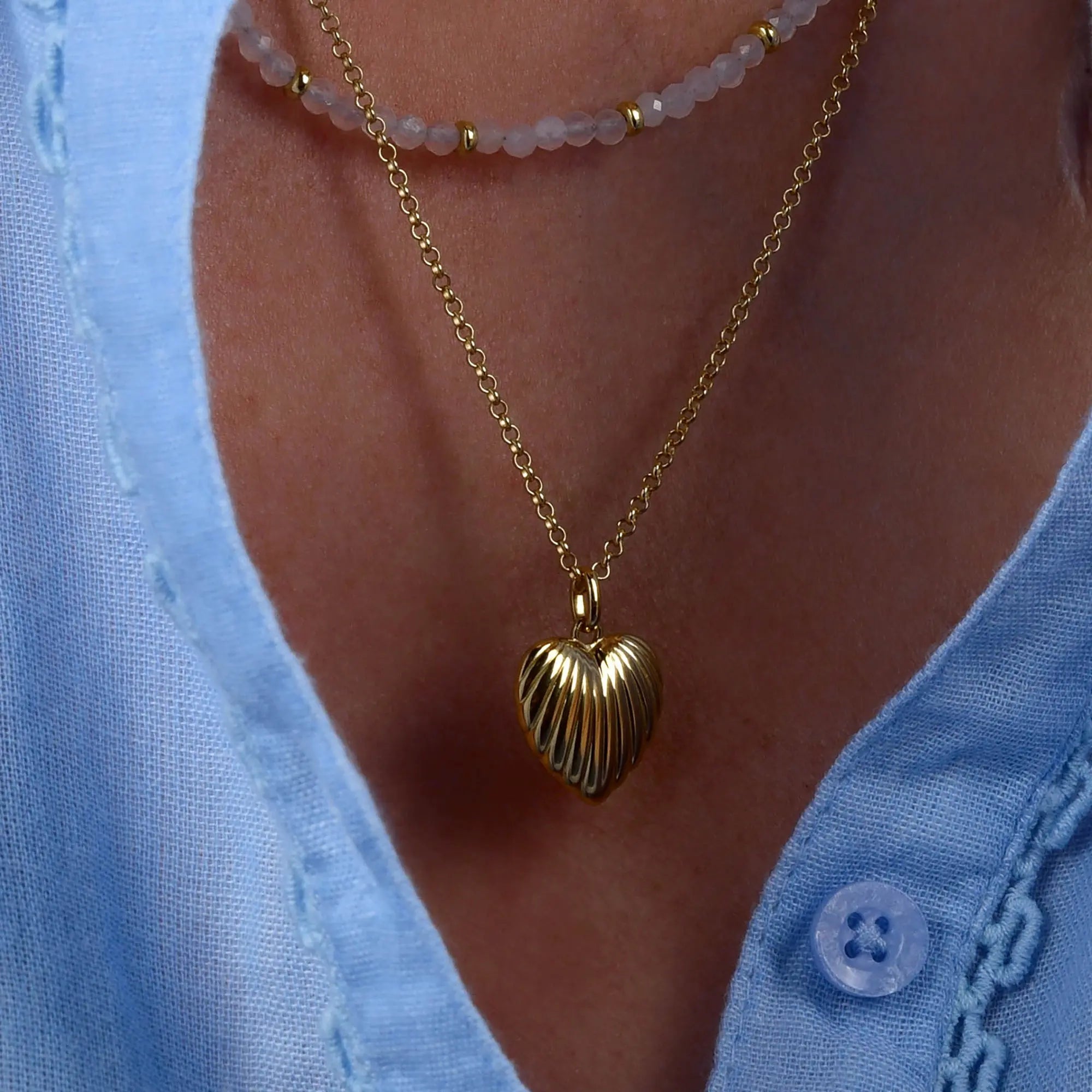 A person wearing a light blue shirt with a gold heart-shaped pendant necklace, styled alongside a beaded choker and the elegant Scallop Heart Necklace.