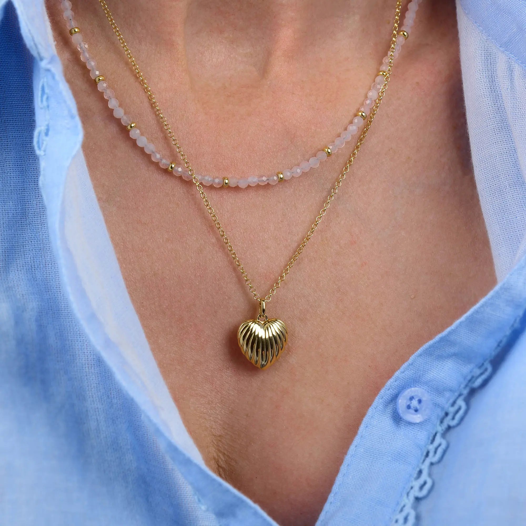 Close-up of a person wearing two necklaces: one is the Rose Quartz Orbit Necklace, a handcrafted piece with pink beads, while the other features a gold heart-shaped pendant. Both are layered over a light blue shirt with embroidery on the collar.