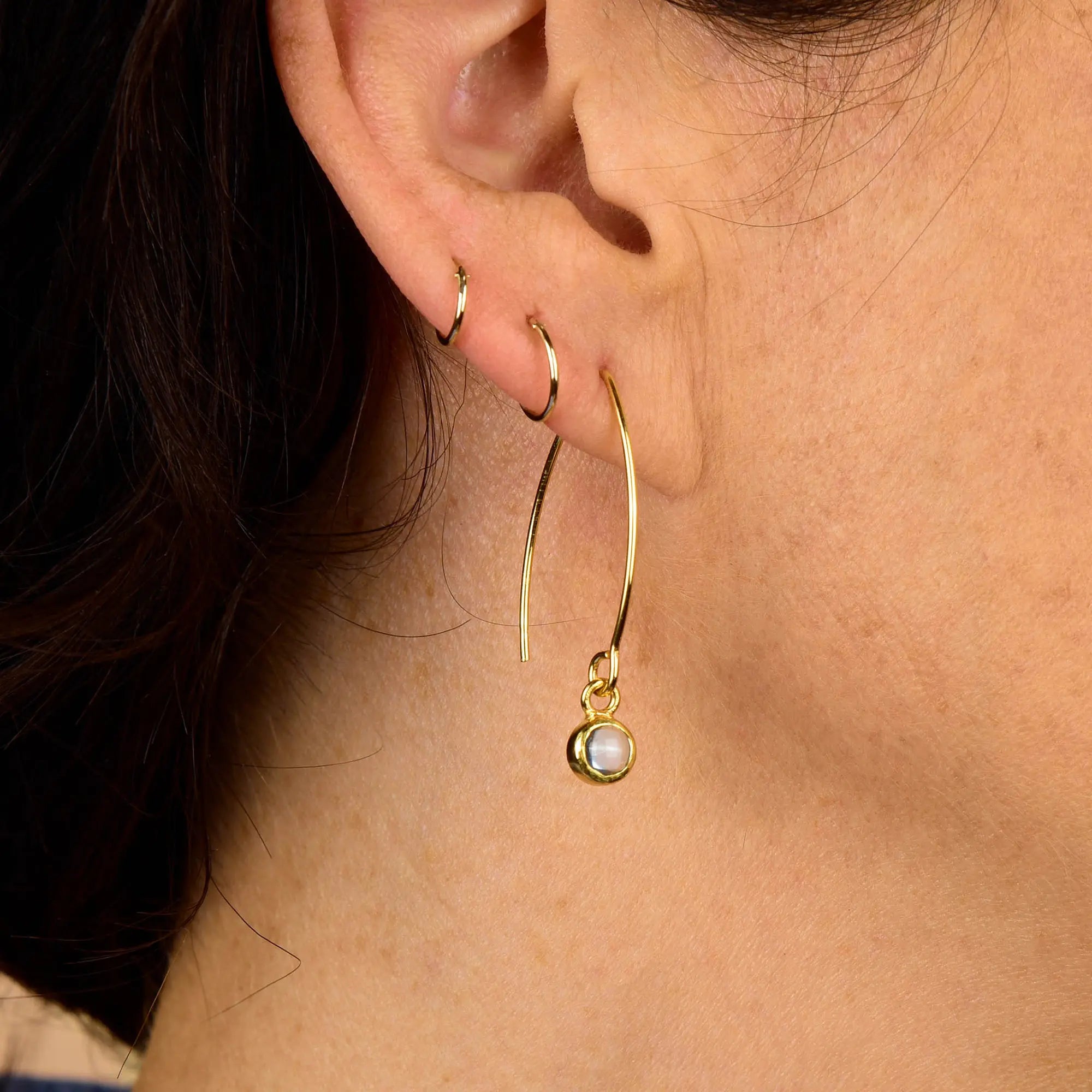 A close-up of a person’s ear wearing three gold earrings: two small hoops and a larger Rose Quartz Gem Dot Arc Earring featuring a dangling pearl charm.