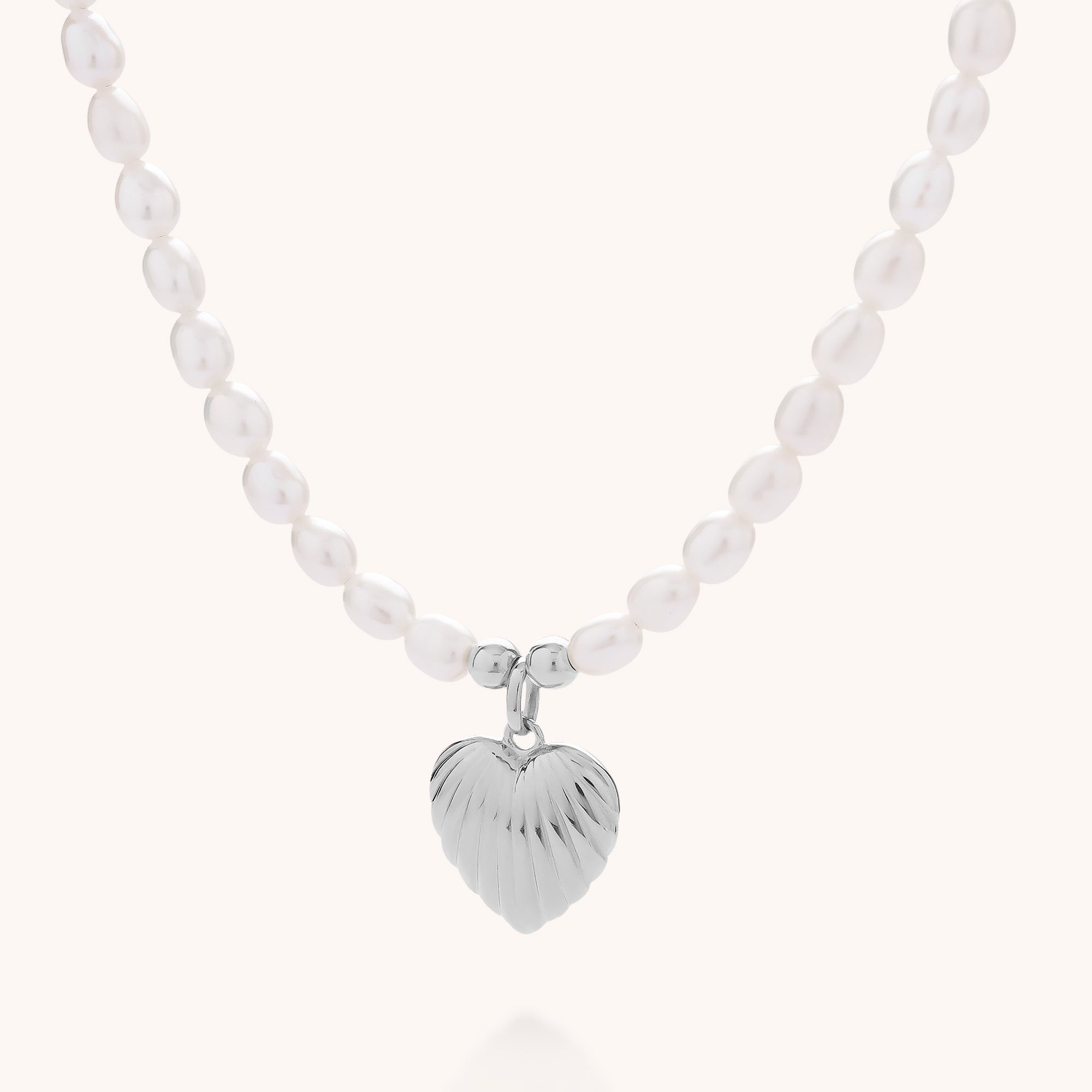 The Scallop Heart Pearl Bead Necklace showcases a silver, textured heart pendant and luminous freshwater pearls. Eco-friendly sterling silver adds charm, making this necklace stand out elegantly against a simple light background.