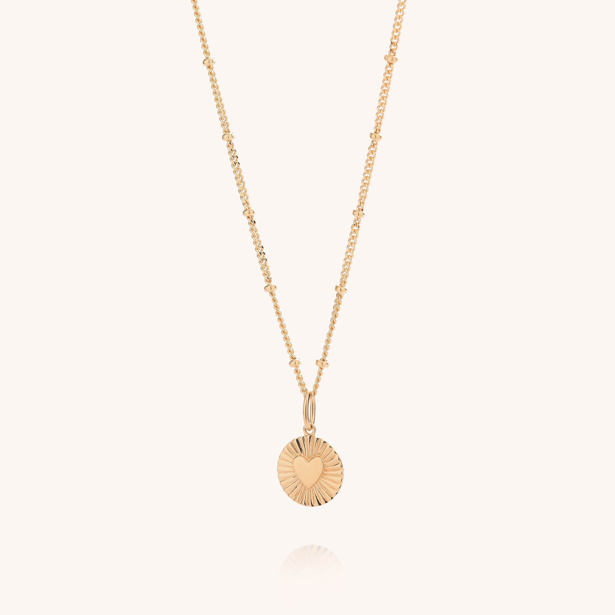 The Shining Heart Necklace Sterling Silver features a gold-plated beaded chain and a round pendant with a central heart design accented by radiating engraved lines. This elegant necklace stands out beautifully against the plain, light-colored background.
