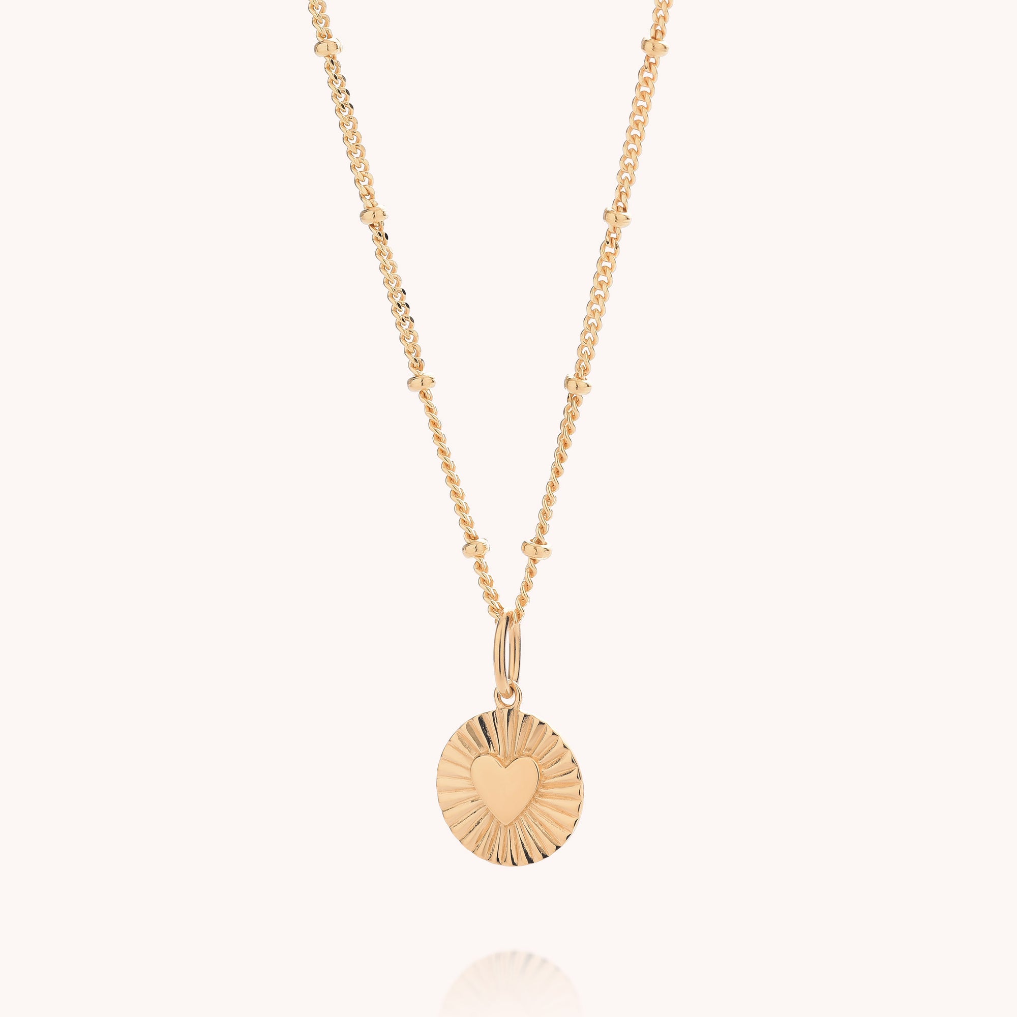 A delicate gold necklace featuring a round sunburst pendant with a heart cutout in the center, suspended from a fine beaded chain against a light cream background.