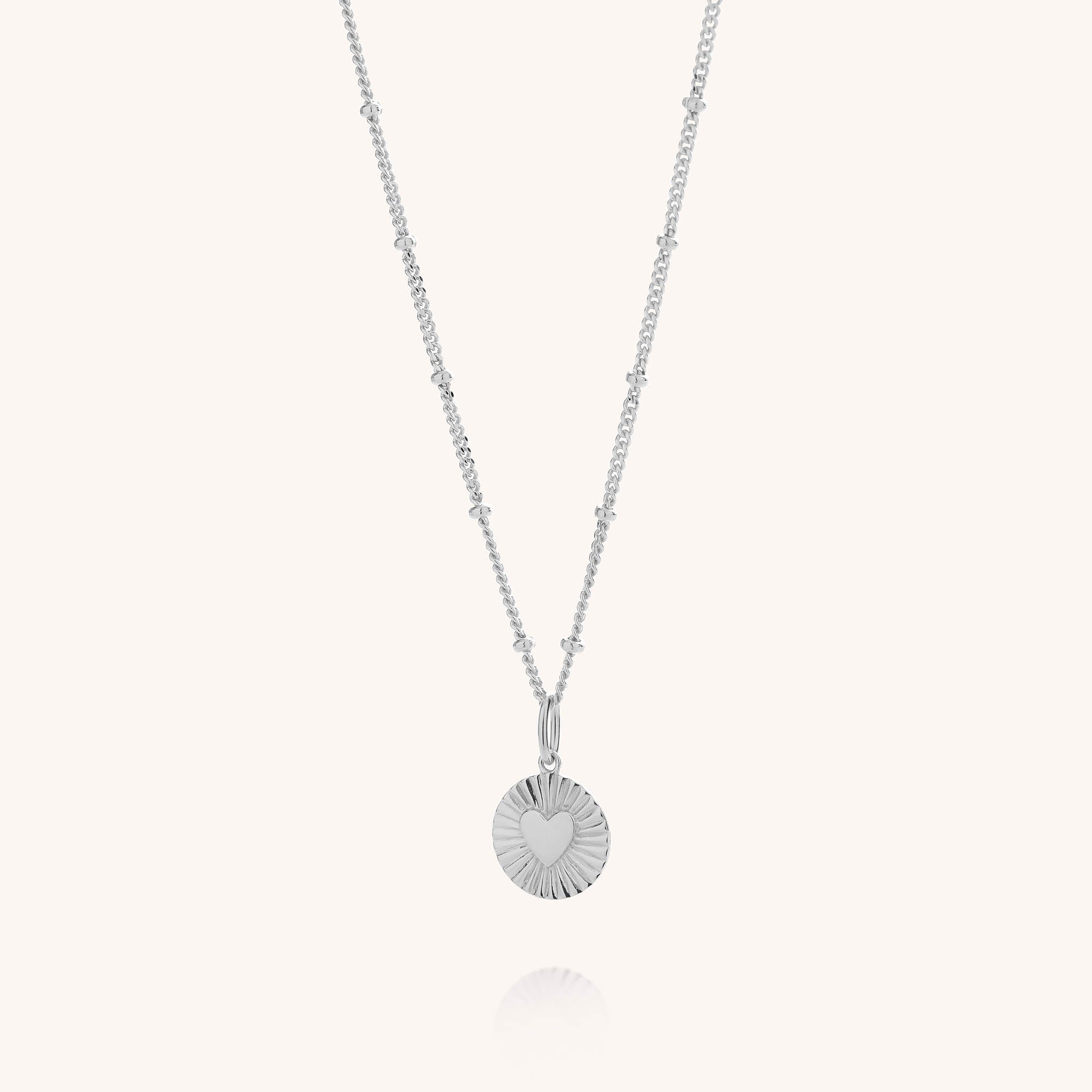 Shining Heart Necklace Sterling Silver featuring a round, ridged pendant with an engraved heart at the center on a sterling silver beaded chain, displayed against a plain light background.