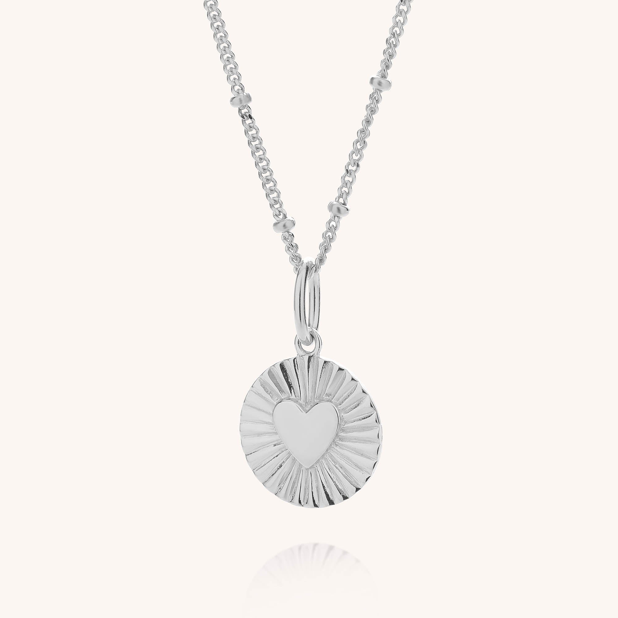 The Shining Heart Necklace Sterling Silver features a round sterling silver pendant with a raised heart in the center, radiant engraved lines, and hangs from a delicate chain adorned with small bead accents.